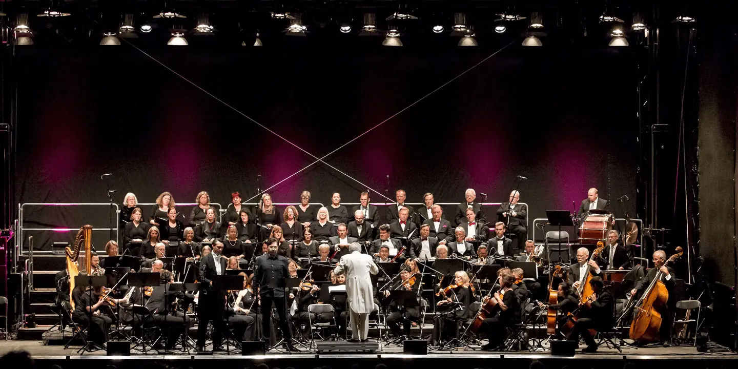 Großes Orchester mit Chor auf Bühne, Dirigent in Weiß leitet Aufführung vor dunklem Hintergrund