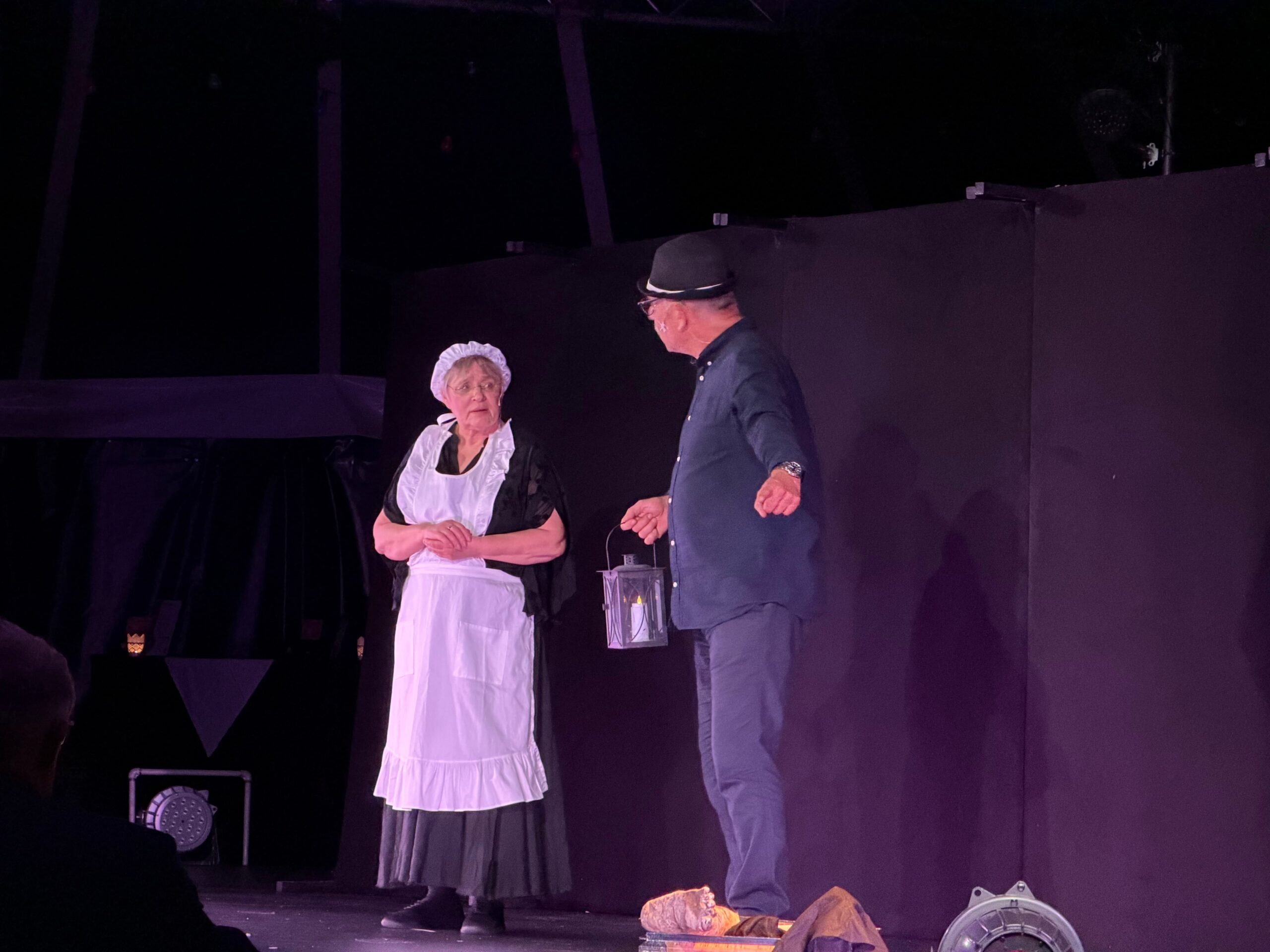 Eine Frau mit schwarzem Kleid und weißer Schürze und Häubchen auf dem Kopf schaut zu einem Mann, ebenfalls in schwarz gekleidet. Er erzählt ihr etwas und hält eine Laterne mit Kerze in der Hand.