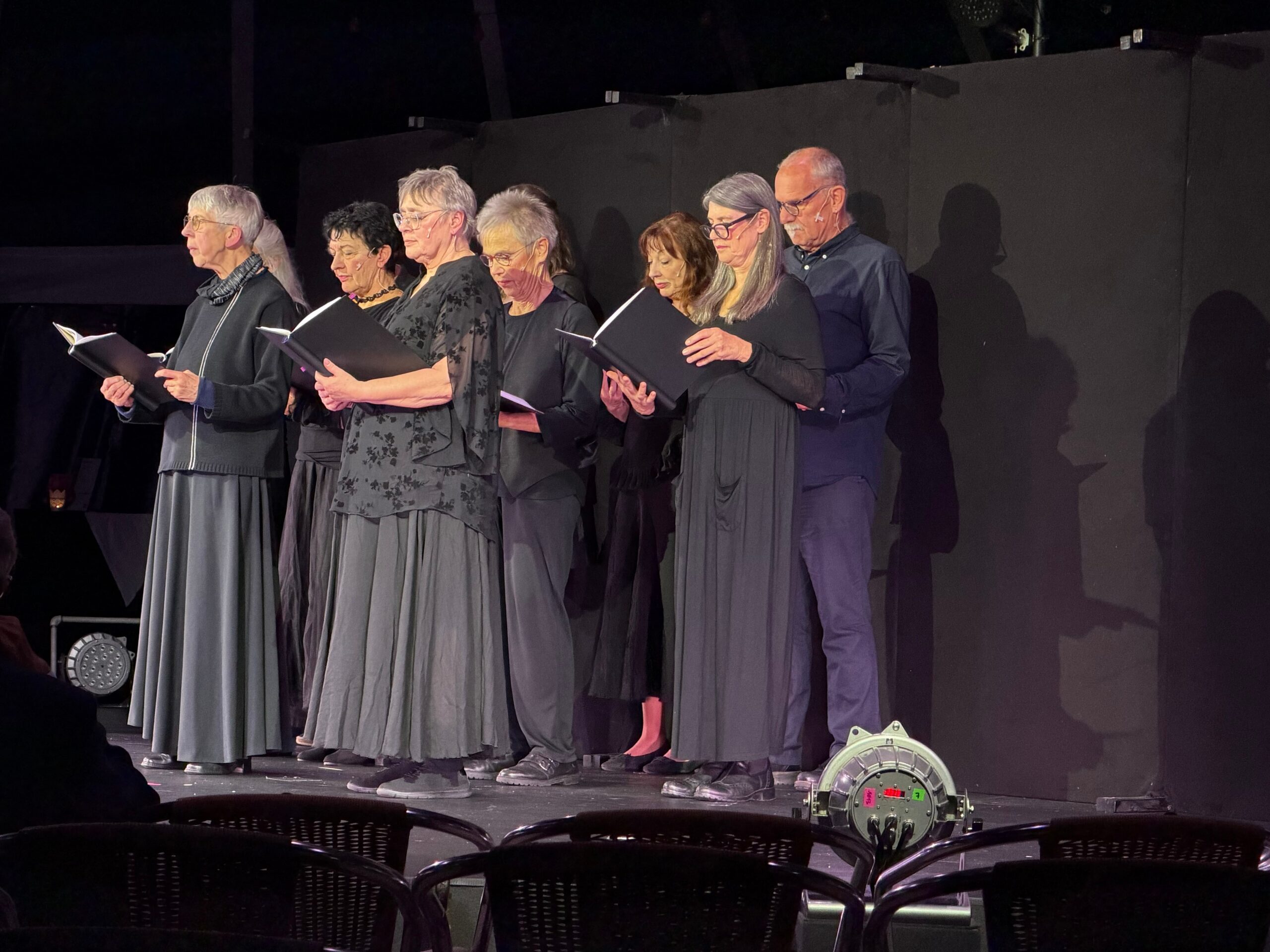8 Seniorinnen und ein Senior, alle in schwarz gekleidet stehen auf einer Bühne und singen aus Büchern.
