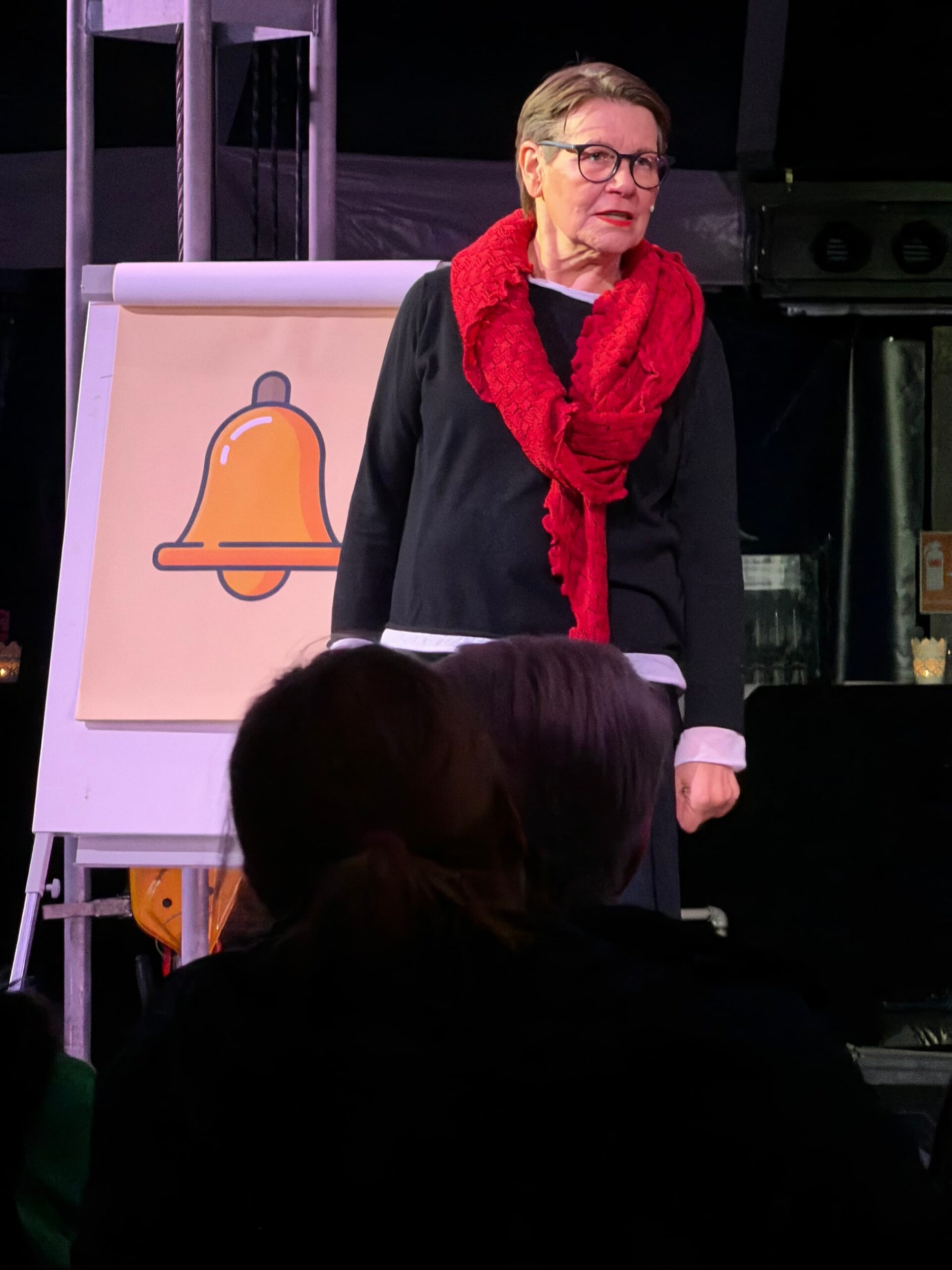 Eine Frau in schwarz gekleidet mit rotem Schal steht vor einer Staffelei mit der Zeichnung einer Glocke und schaut ins Publikum.