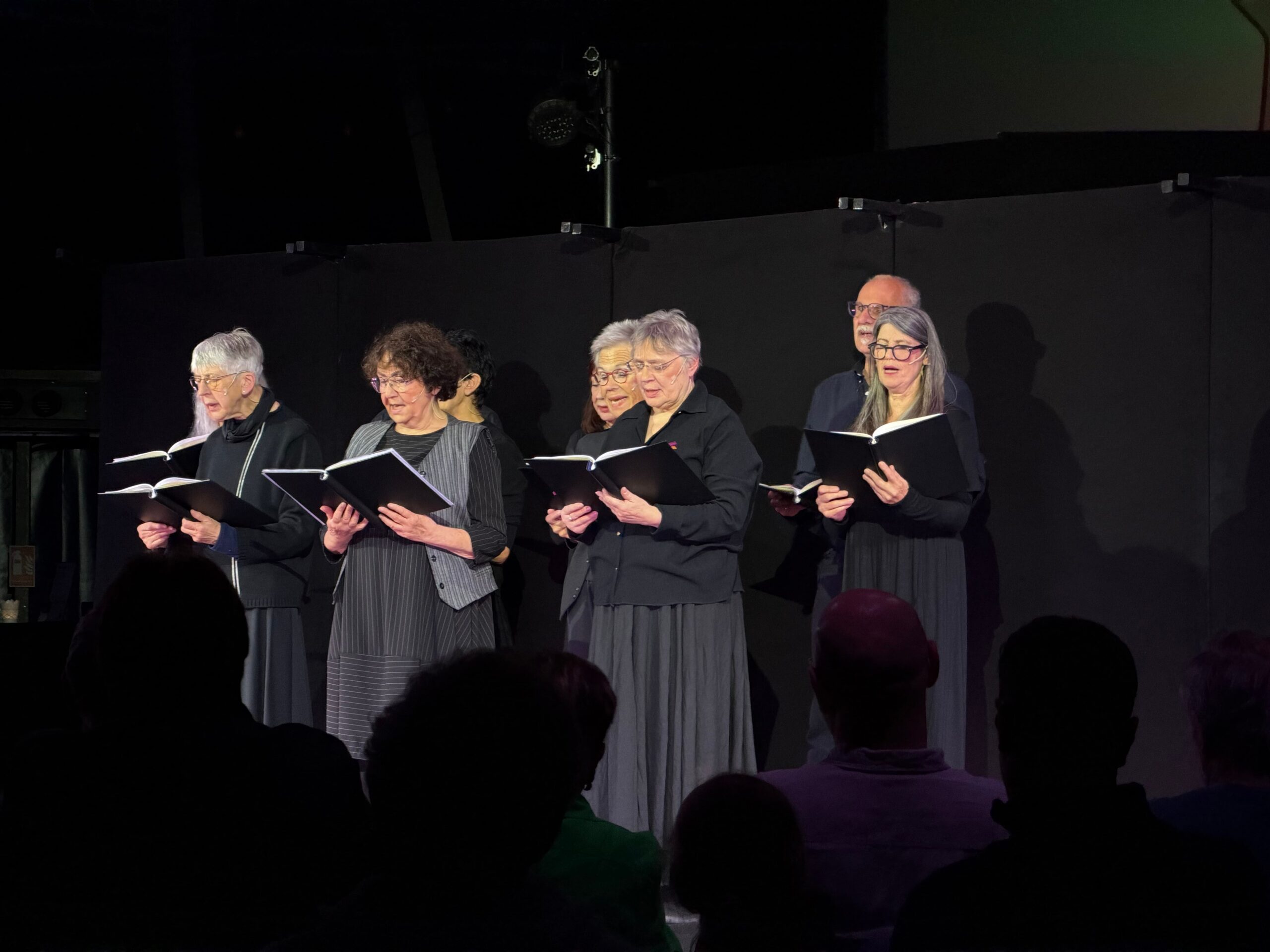 8 Seniorinnen und ein Senior, alle in schwarz gekleidet stehen auf einer Bühne und singen aus Büchern.