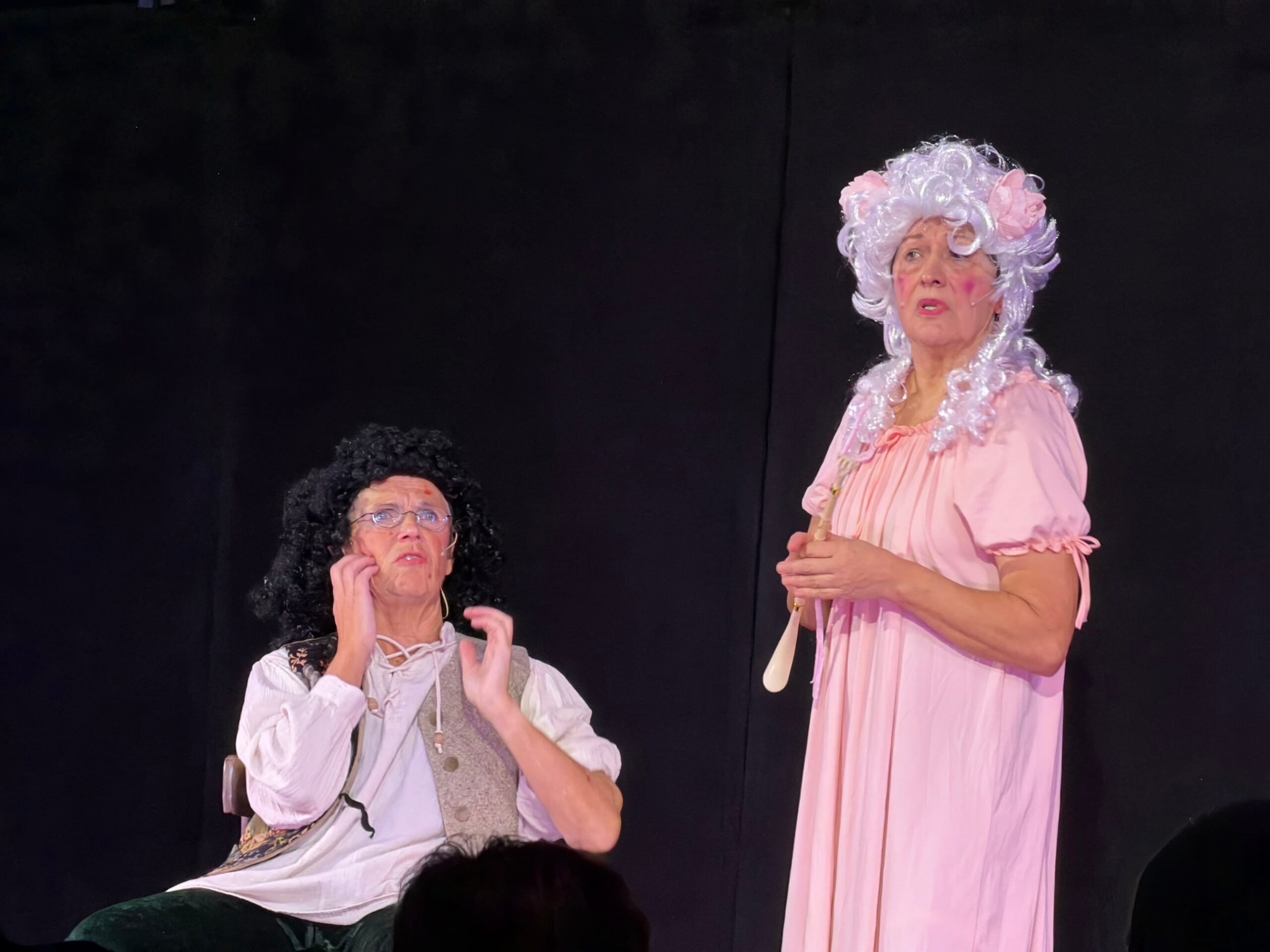 Eine ältere Dame im Kostüm mit weißen, langen Haaren und einem rosafarbenen, langen Kleid steht links. Rechts ist ein älterer Herr ebenfalls im Kostüm, mit schwarzen, langen Haaren, weißem Hemd und brauner Weste auf einem Stuhl sitzend. Beide sind im barocken stil gekleidet und unterhalten sich.