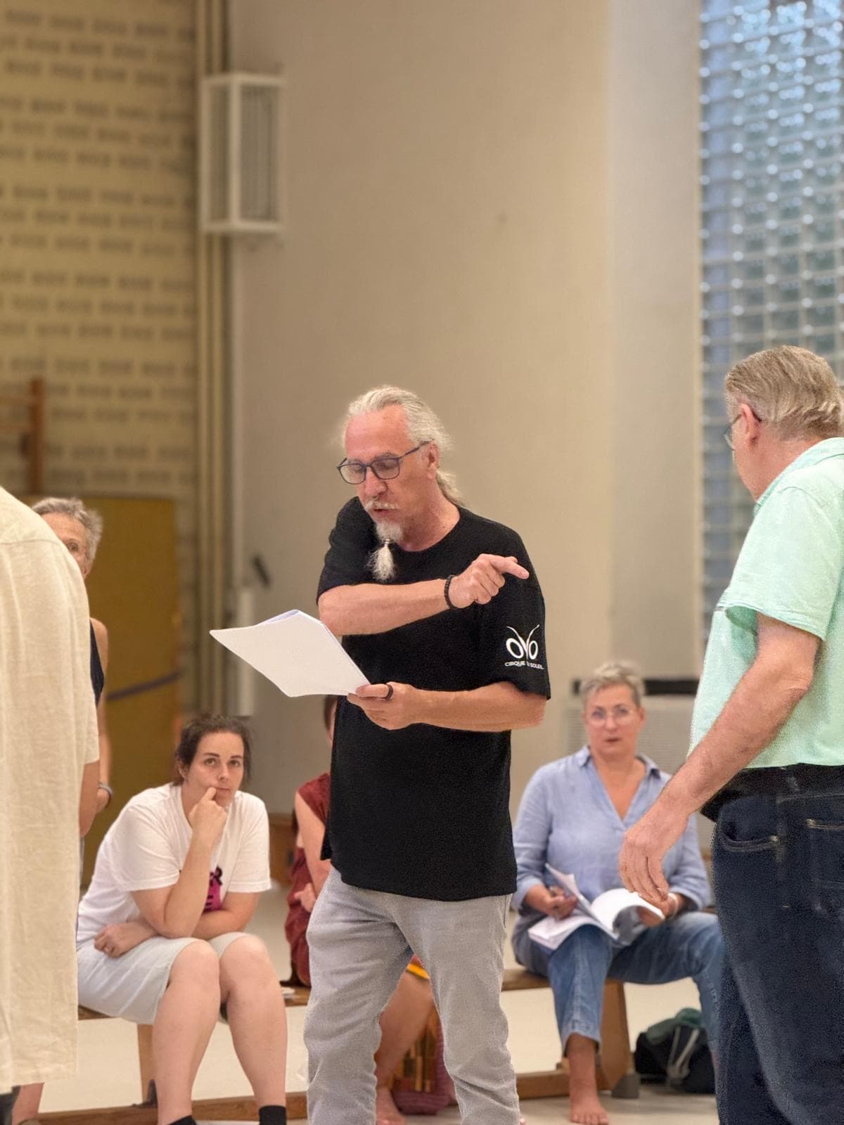 Theaterprobe in einer Halle. Viele Menschen sitzen auf Bänken, ein Mann erklärt in der Mitte etwas.