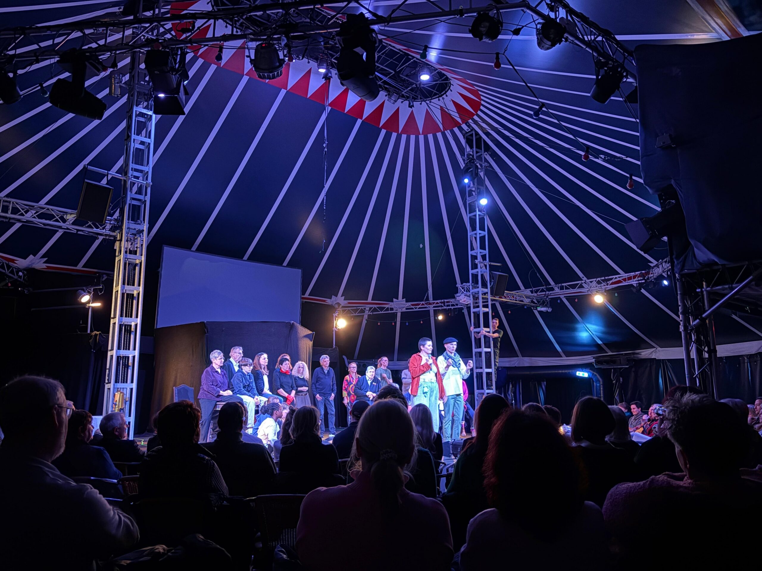 Ein Zirkuszelt, in der Mitte eine Bühne, auf der viele Schauspieler stehen.