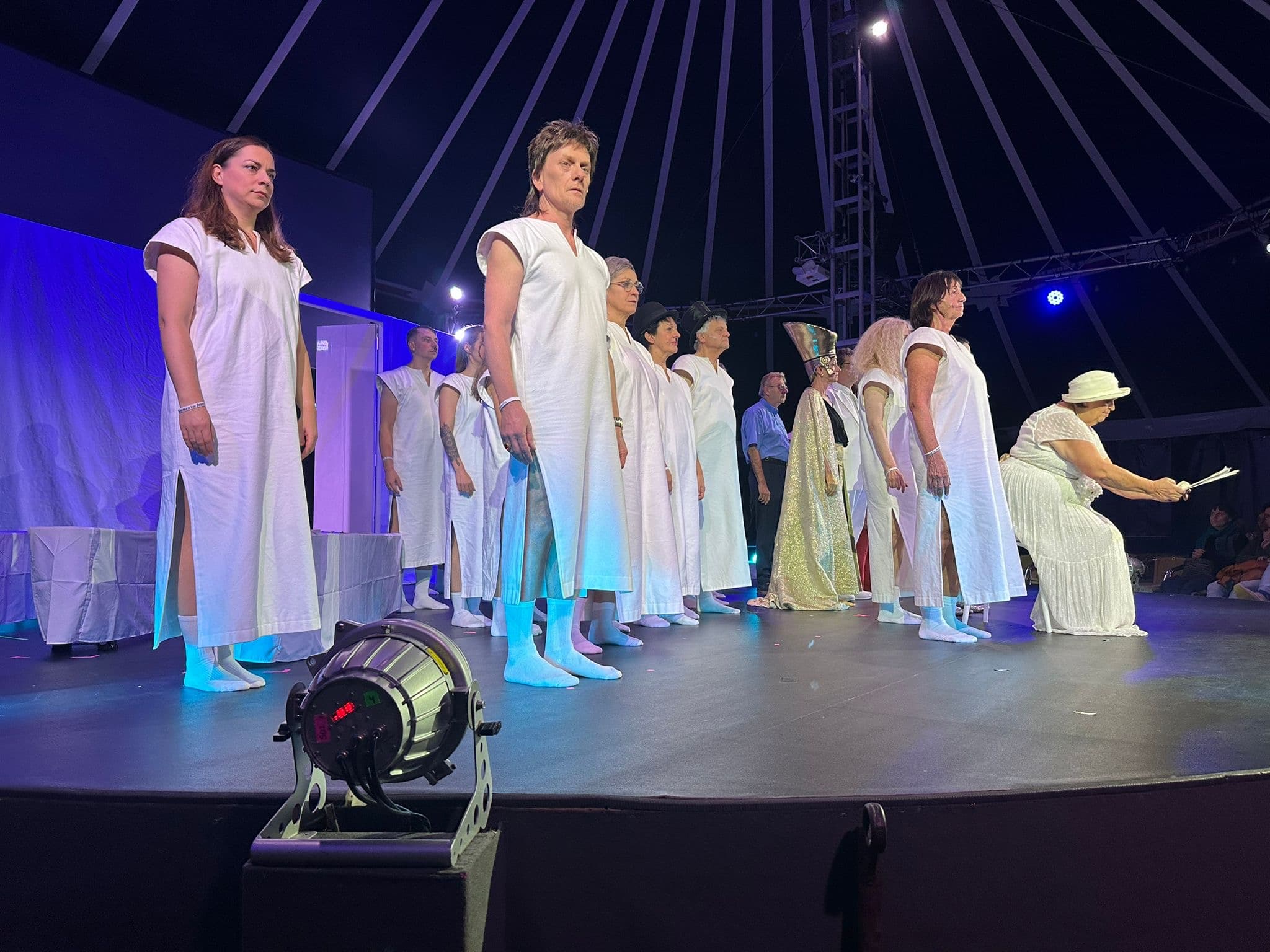 Viele Schauspielerinnen und Schauspieler stehen auf der Bühne. Alle tragen weiße Gewänder, ein ägyptischer Pharao in Gold steht ebenfalls dabei. Eine Frau in Weiß gekleidet mit weißem Hut sitzt auf einem Stuhl ganz vorne und hält Dokumente in der Hand.