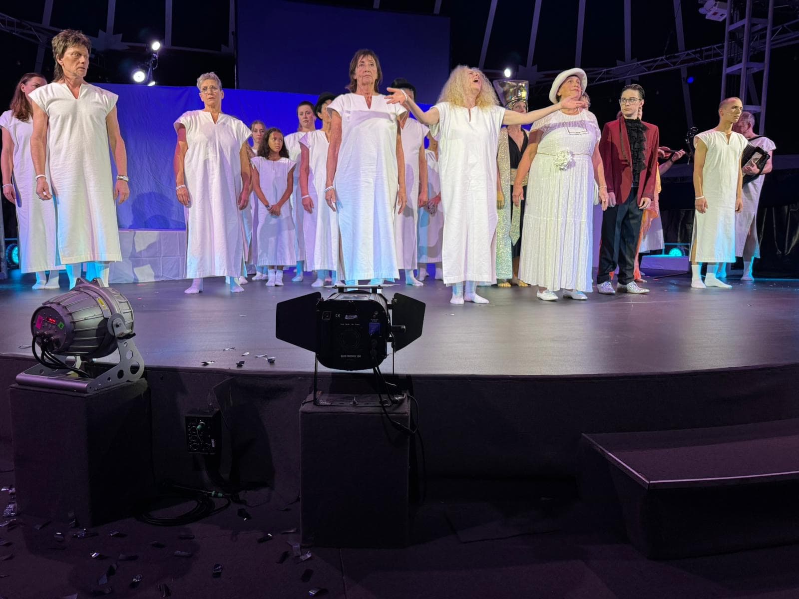 Viele Menschen in weißen Nachthemden stehen auf der Bühne. Vorne in der Mitte steht eine Frau mit langen blonden Haaren, die den Kopf zur Decke hebt und etwas klagt.