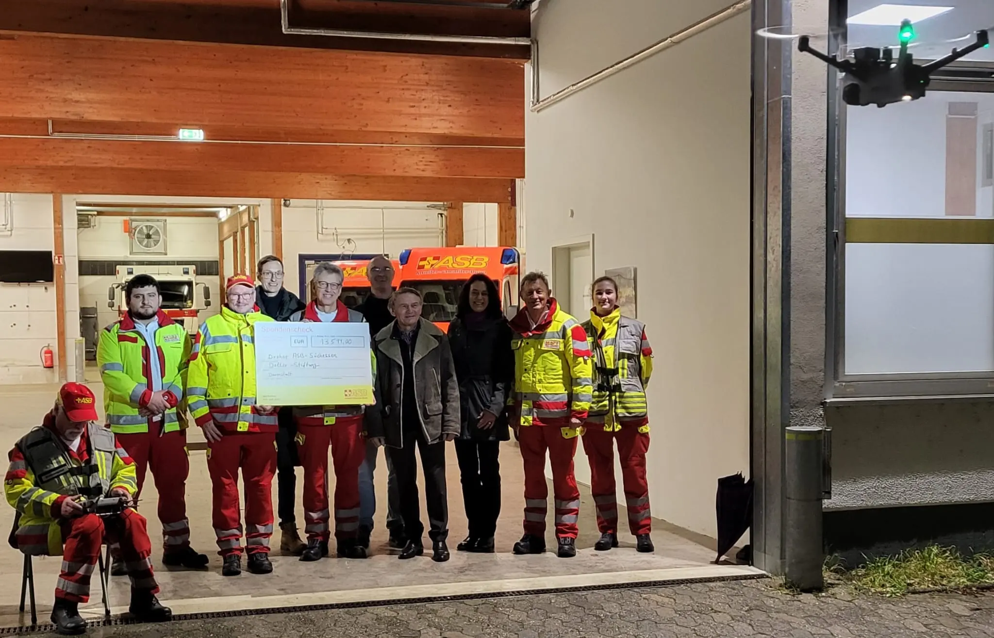 Eine Gruppe von Rettungssanitätern in gelben und roten Uniformen steht vor einem Gebäude, einige halten einen großen Scheck. Zwei Personen in Zivilkleidung stehen in der Mitte. Eine Drohne schwebt in der Nähe auf der rechten Seite.