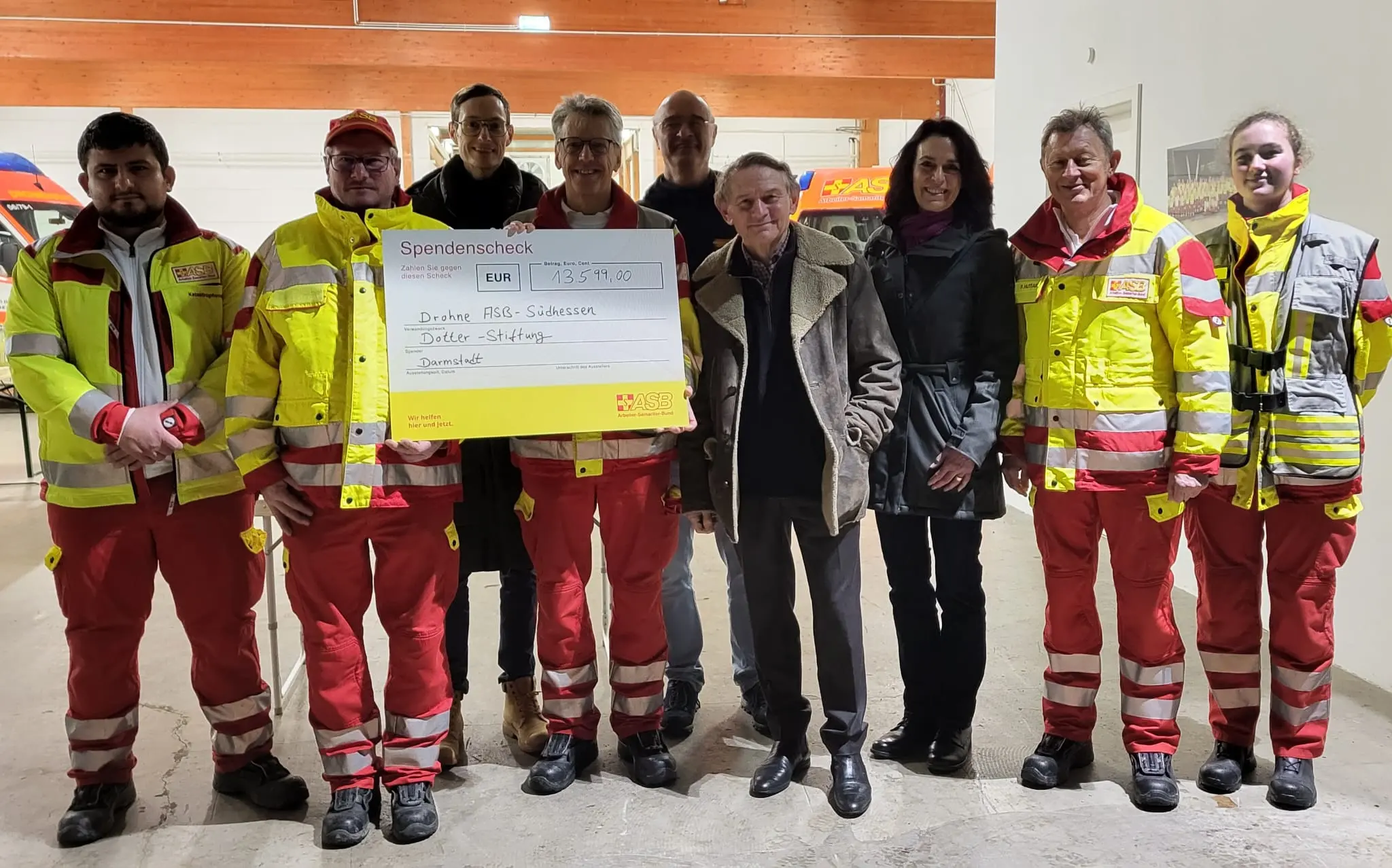 Eine Gruppe von Personen, darunter mehrere in roten und gelben Rettungsdienstuniformen, steht in einem Haus und hält einen großen Spendenscheck in der Hand. Andere in der Gruppe tragen dunkle Kleidung, und alle schauen in die Kamera und lächeln.