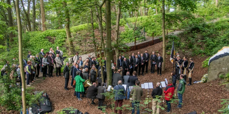 Eine große Gruppe von Menschen versammelt sich in einem bewaldeten Gebiet, einige förmlich in Anzügen, andere leger gekleidet. Im Vordergrund sitzen Musiker, und die Szene scheint eine Zeremonie oder eine Veranstaltung im Freien unter Bäumen zu sein.