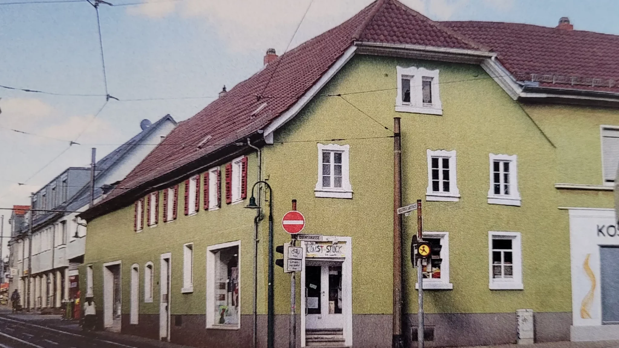 Ein grünes, zweistöckiges Eckgebäude mit rotem Ziegeldach hat weiß gestrichene Fenster und einen Ladeneingang an der Ecke. Verkehrsschilder und Straßenlaternen sind auf dem Gehweg neben dem Gebäude zu sehen.