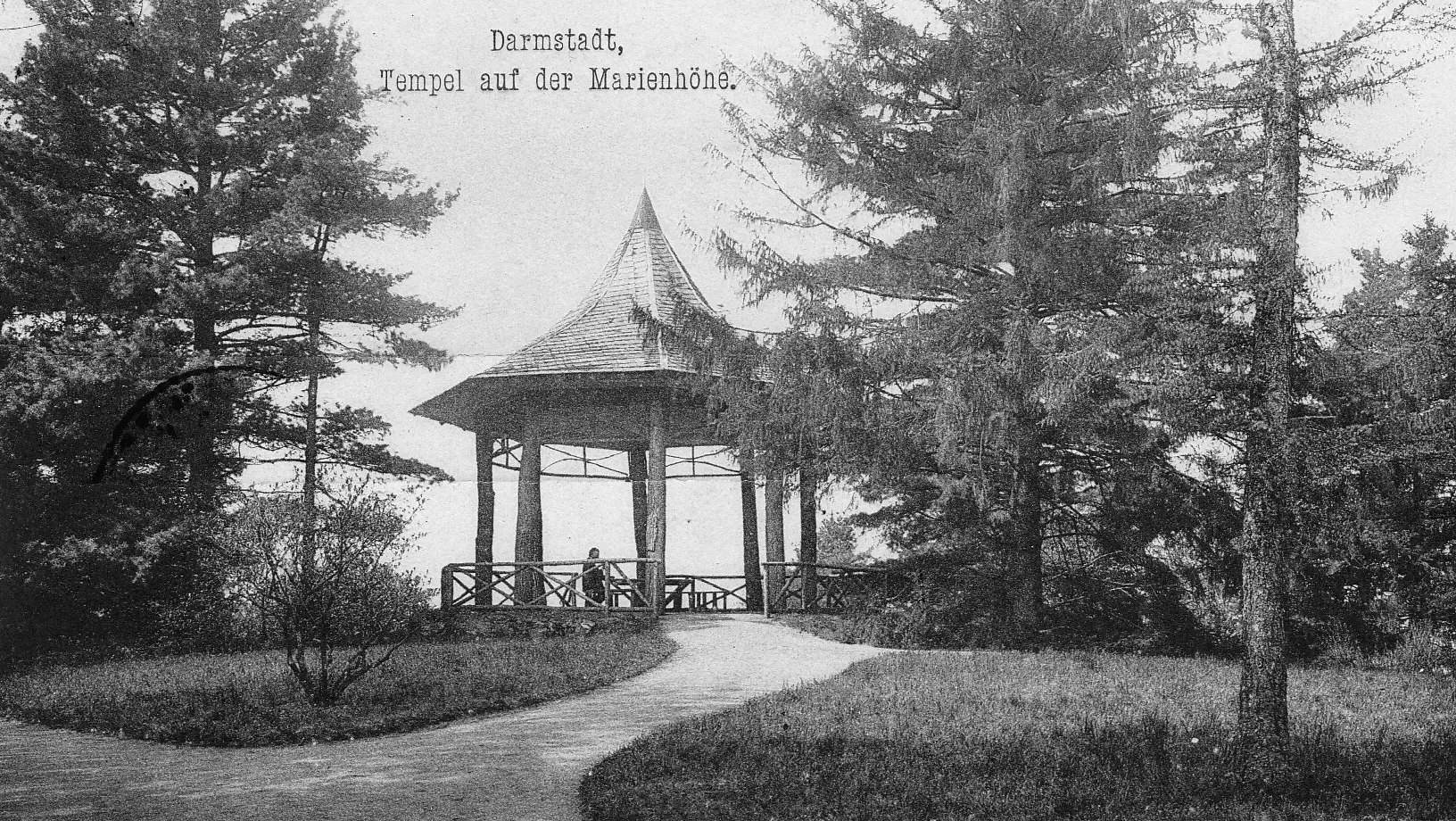 Schwarz-Weiß-Foto eines sechseckigen Holzpavillons mit Spitzdach, umgeben von hohen Bäumen. Darin sitzt eine Person. Der Text lautet: "Darmstadt, Tempel auf der Marienhöhe". Wege führen zu dem Bauwerk.