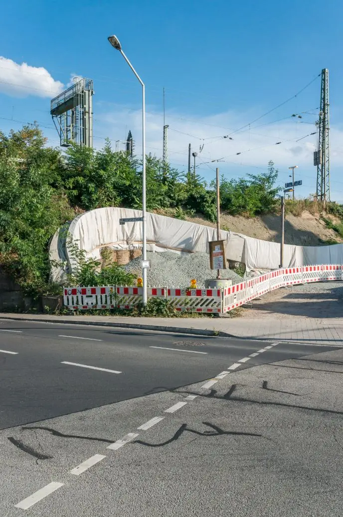 Eine Baustelle ist mit roten und weißen Zäunen neben einer Straße und einem Gehweg abgesperrt. Zu sehen sind Schotterhaufen und Baumaterialien, im Hintergrund sind Grünpflanzen und Strommasten unter blauem Himmel zu sehen.