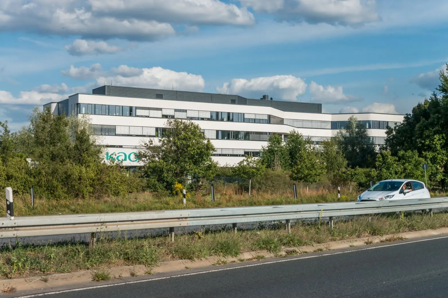Ein modernes, weißes Bürogebäude, das teilweise von Bäumen verdeckt wird, steht hinter einer Straße, an der ein weißes Auto vorbeifährt, unter einem blauen Himmel mit vereinzelten Wolken. Auf dem Gebäude ist das Logo "kao." zu sehen.