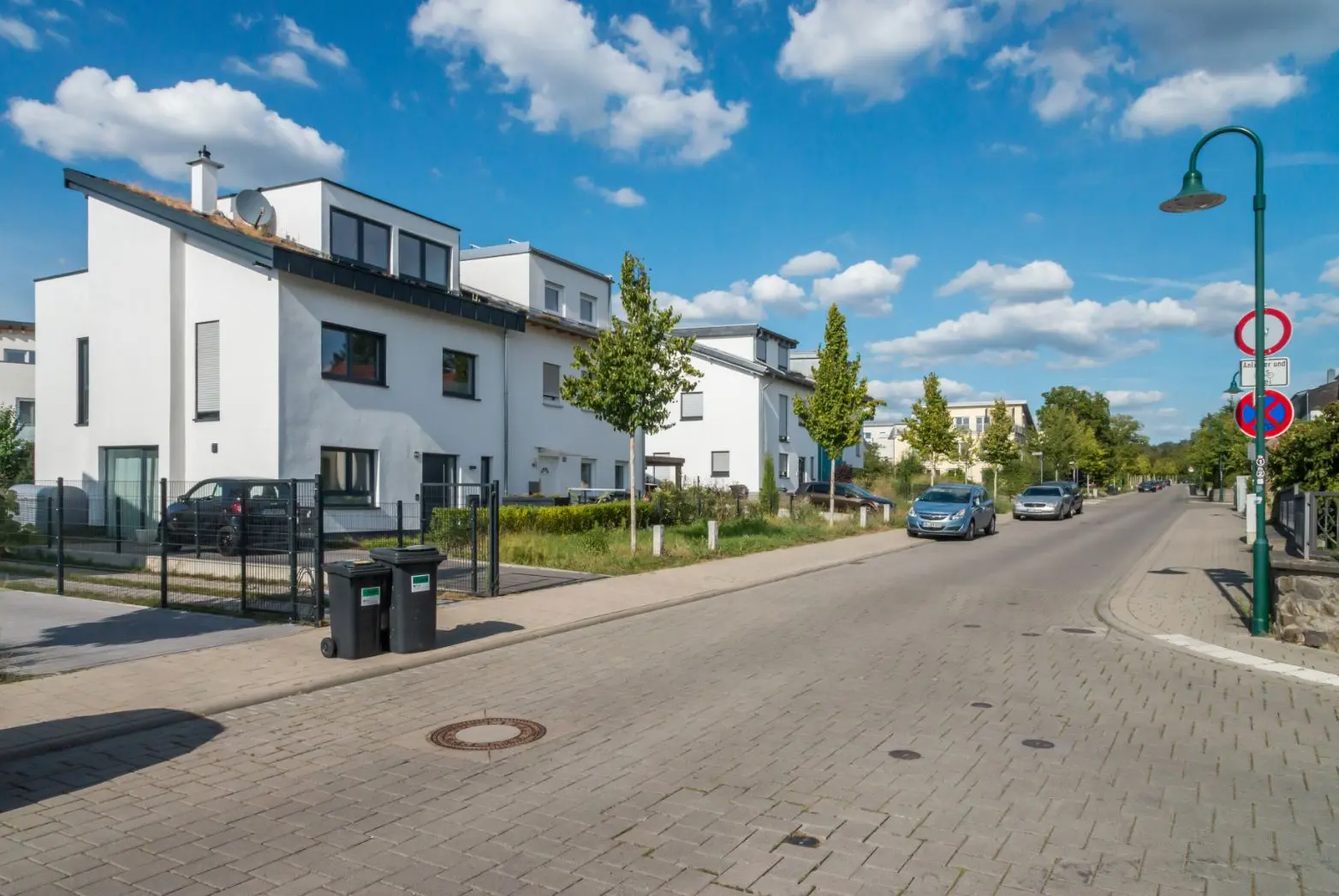 Eine ruhige Vorstadtstraße mit modernen weißen Häusern, grünen Bäumen, geparkten Autos, Mülleimern auf dem Gehweg und einem blauen Himmel mit vereinzelten Wolken. Auf der rechten Seite der Straße sind Straßenlaternen und Verkehrsschilder zu sehen.