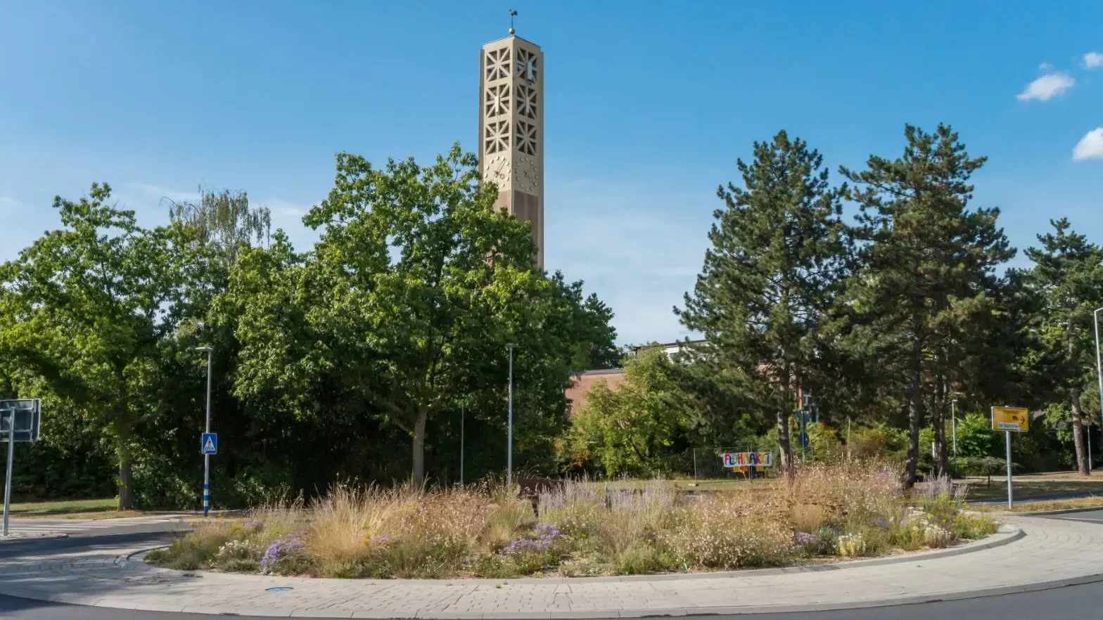 Ein Kreisverkehr mit Wildblumen und hohem Gras in der Mitte, umgeben von Bäumen. Ein hoher, beiger Kirchturm mit geometrischen Mustern erhebt sich über dem Grün unter einem klaren blauen Himmel.