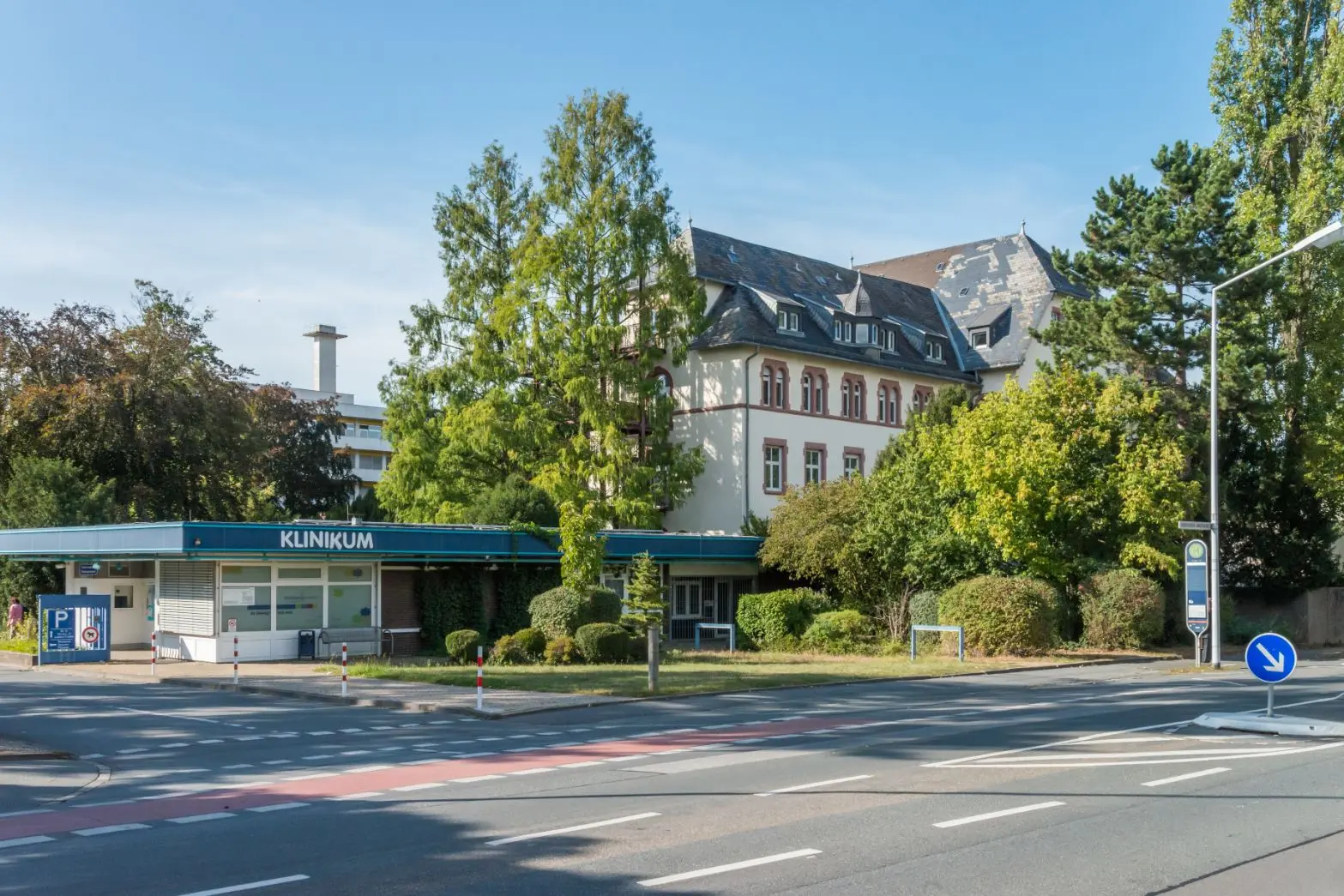 Ein Krankenhausgebäude mit der Aufschrift Klinikum steht an einem sonnigen Tag hinter Bäumen und Büschen, im Vordergrund sind ein Zebrastreifen und Straßenschilder zu sehen.