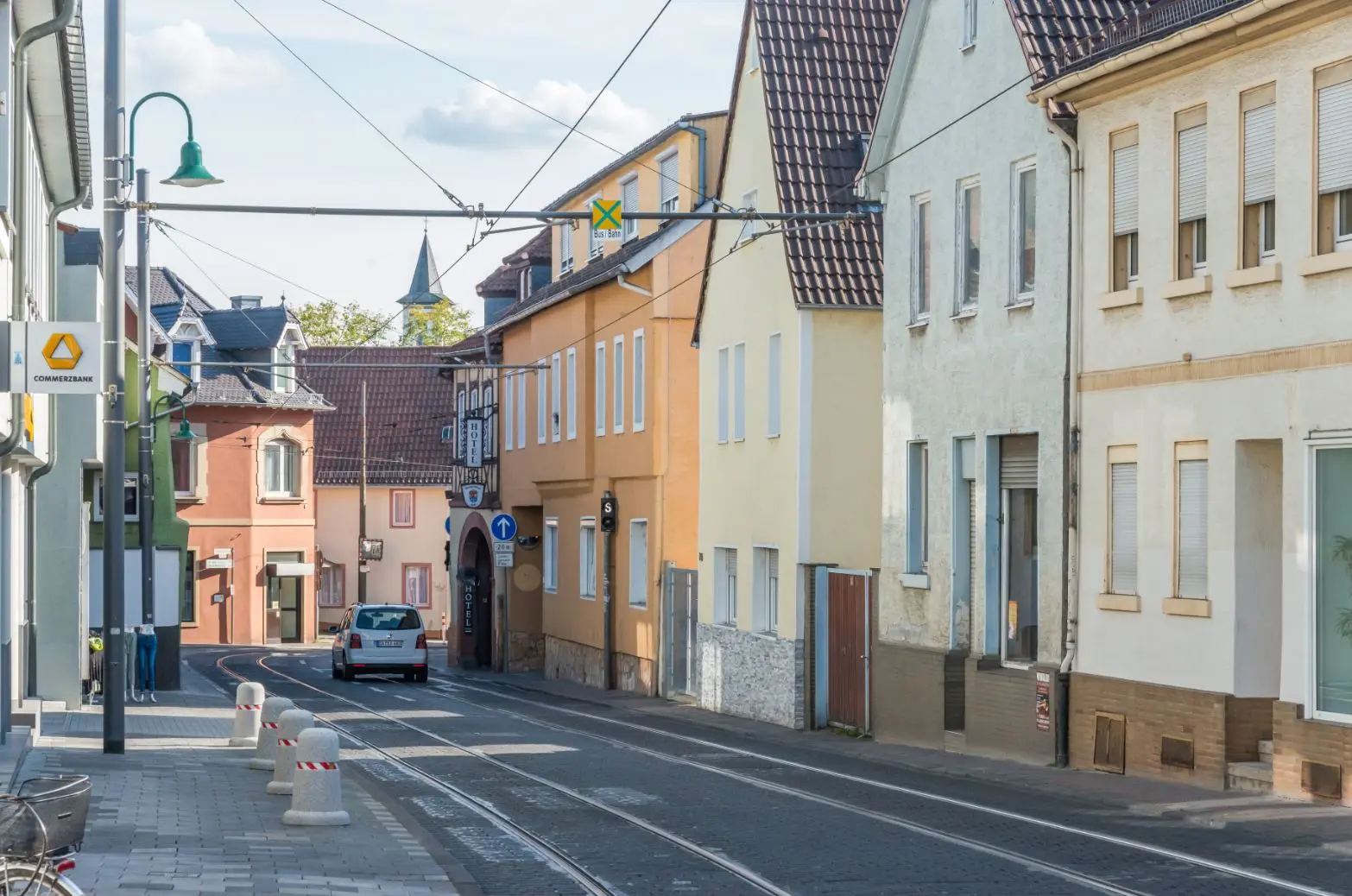 Eine ruhige Straße in einer europäischen Stadt mit pastellfarbenen Gebäuden, ein paar geparkten Autos, Straßenbahnschienen und Oberleitungen. Die Straße ist leer und wird von Bürgersteigen und Straßenlaternen gesäumt.