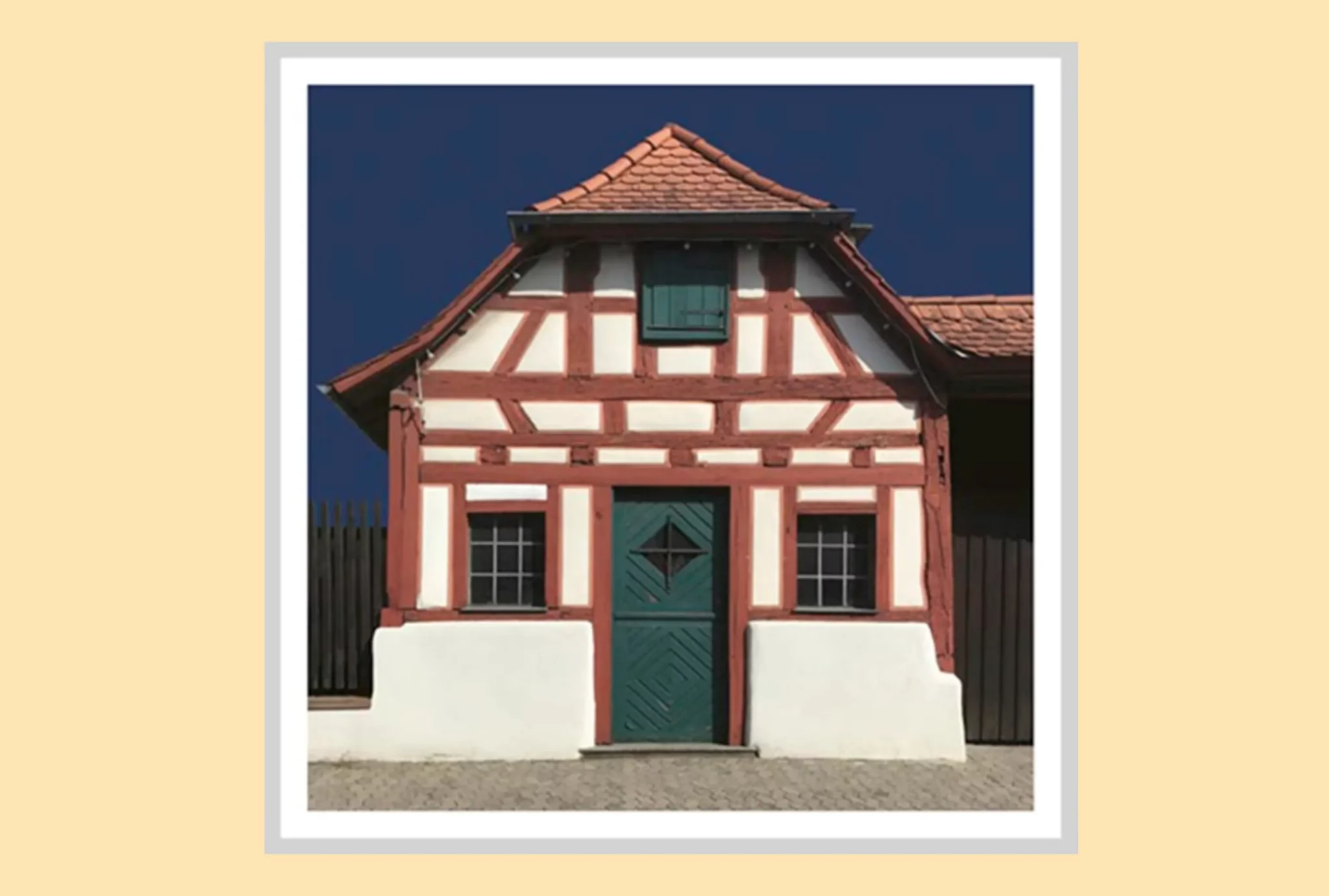 Ein kleines, traditionelles Fachwerkhaus mit roten Holzbalken, weißen Wänden, einer dunkelgrünen Tür und einem Fenster mit Fensterläden vor einem tiefblauen Himmel.