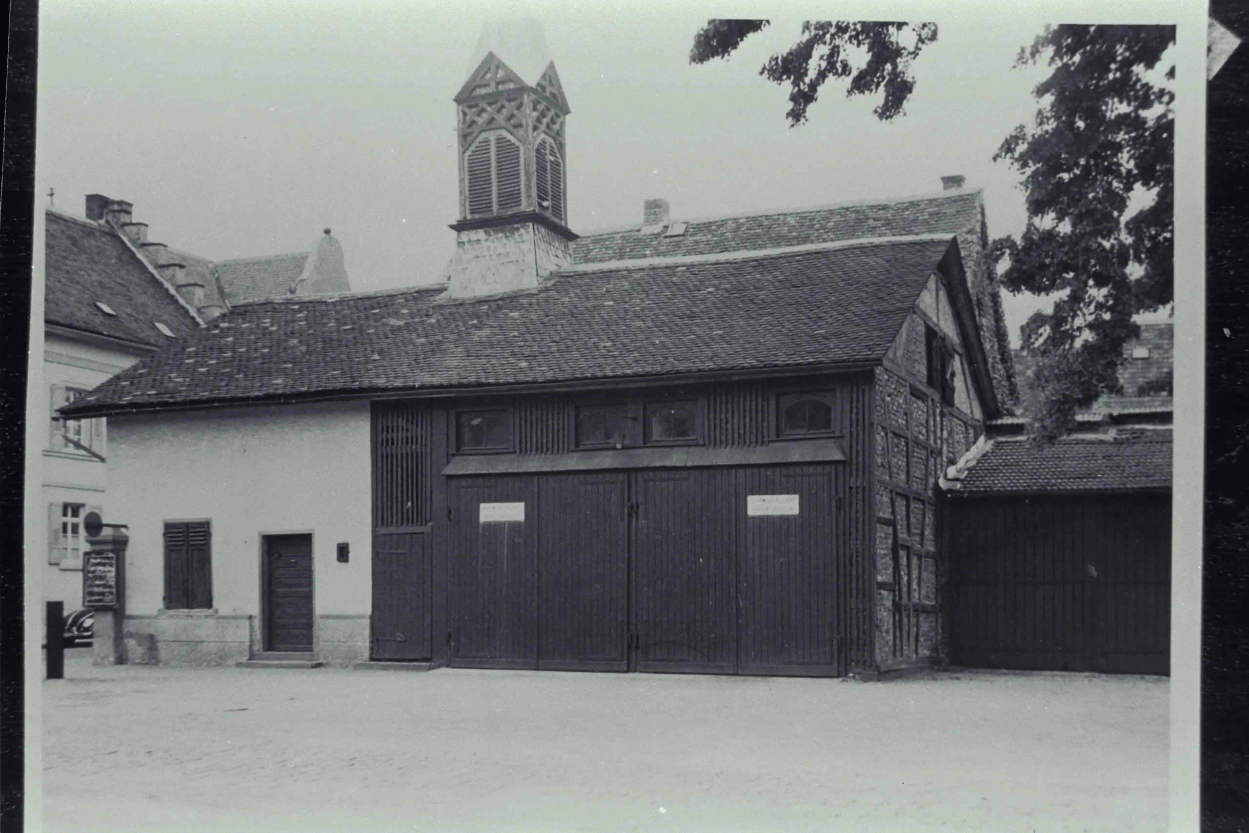 Schwarz-Weiß-Foto eines Gebäudes mit großen Holztüren, einem kleinen Uhrturm und einem Ziegeldach. Auf der rechten Seite sind Bäume zu sehen, und links ist ein Wegweiser zu erkennen.
