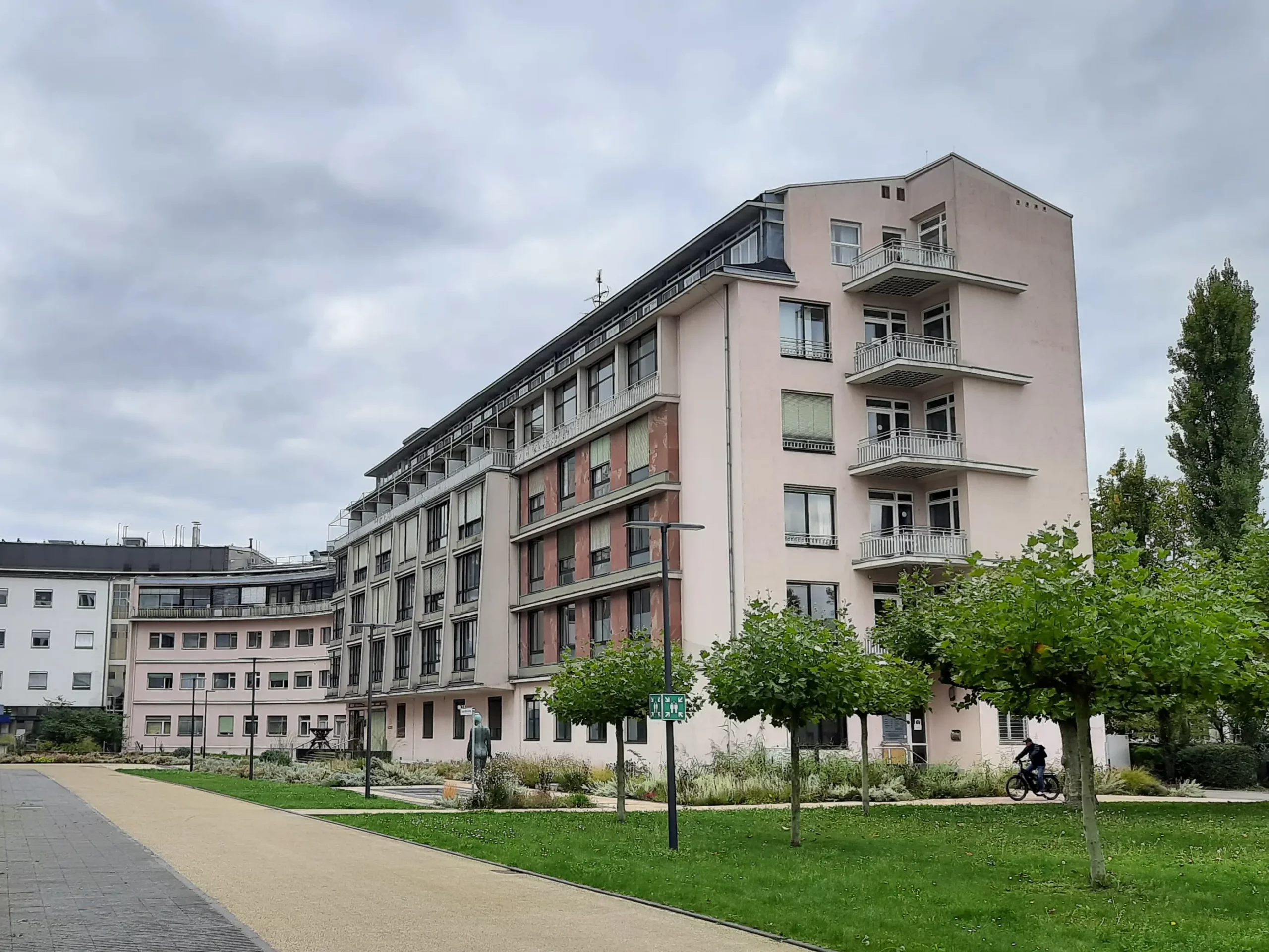 Ein mehrstöckiges, rosa-weißes Wohnhaus mit Balkonen, das neben einer grünen Rasenfläche mit jungen Bäumen unter einem bewölkten Himmel steht. Eine Person fährt mit dem Fahrrad in der Nähe des Gebäudes.
