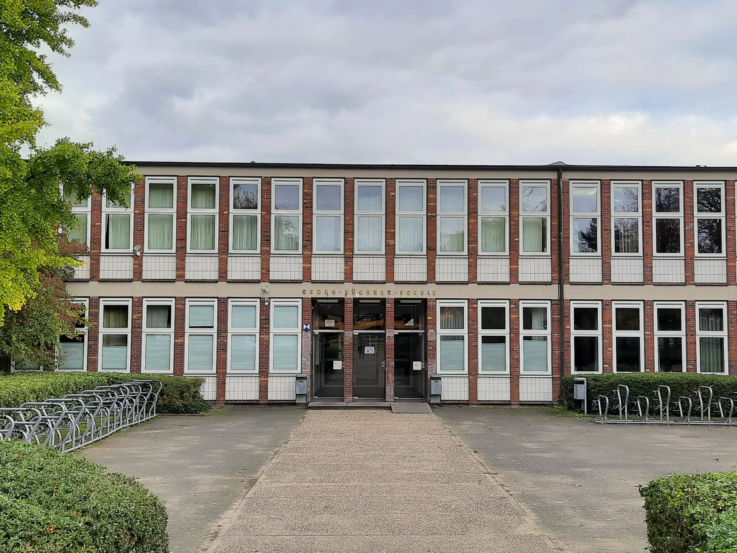 Ein zweistöckiges Schulgebäude mit großen Fenstern und einem zentralen Eingang, umgeben von Büschen, Bäumen und einem Fahrradständer auf der linken Seite unter bewölktem Himmel.