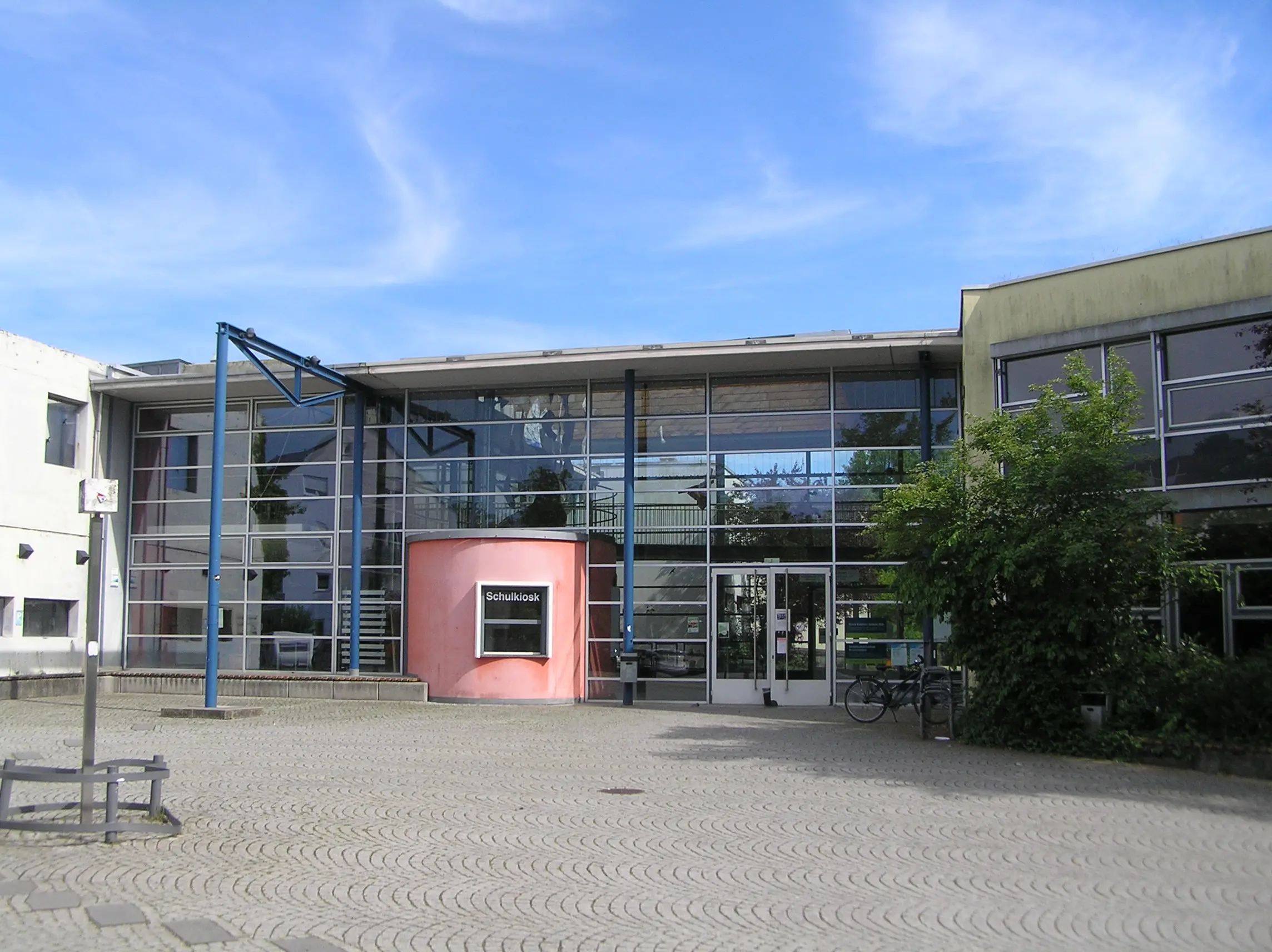 Ein modernes Schulgebäude mit großen Glasfenstern, einem rosafarbenen, geschwungenen Eingangsbereich und einem Schild mit der Aufschrift „Schulbibliothek“. Vor dem Eingang stehen Fahrräder, und an einem sonnigen Tag sind draußen Bäume zu sehen.