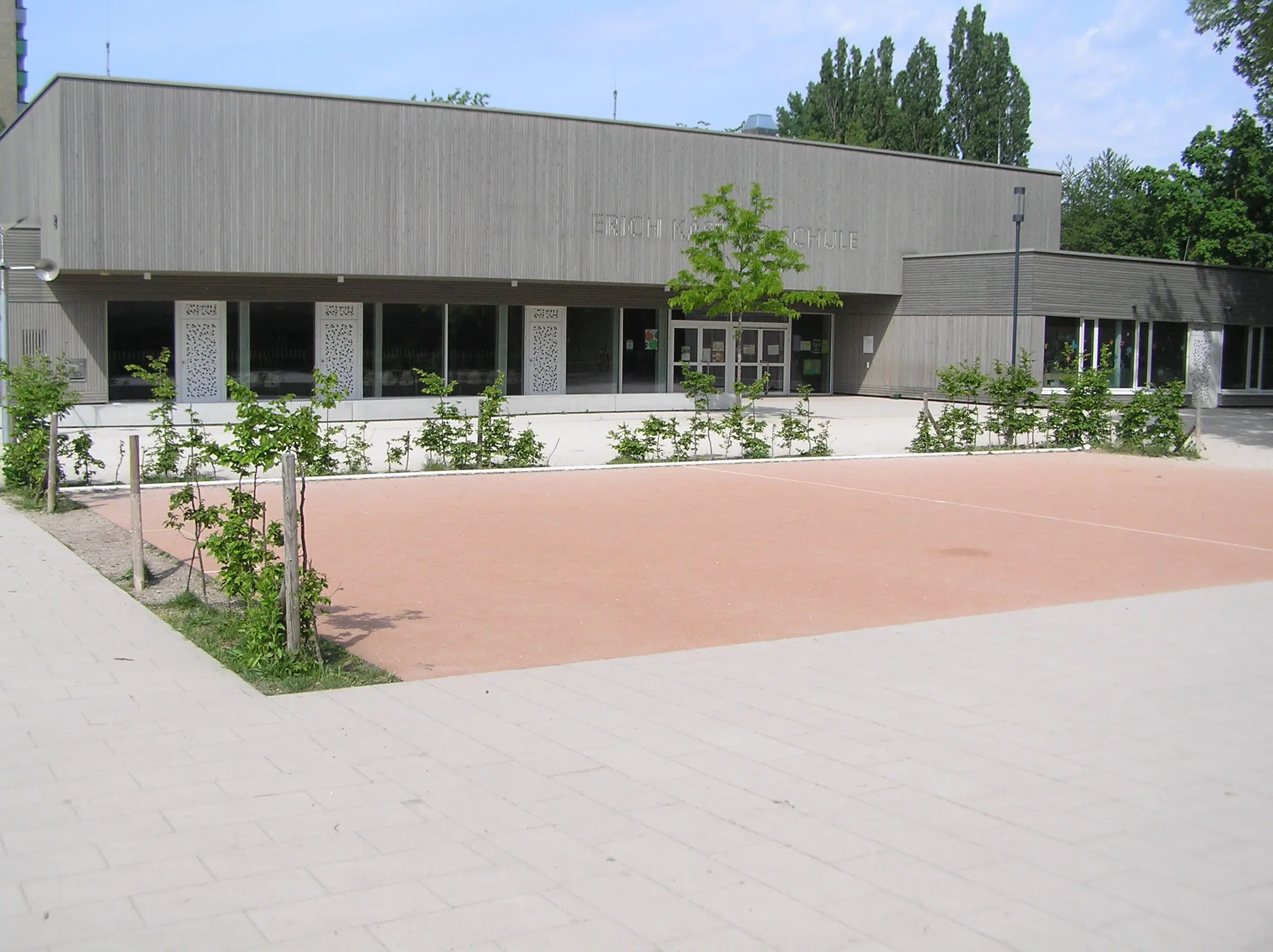 Hinter einem umzäunten, leeren Sportplatz und einer Reihe kleiner grüner Büsche steht ein modernes, rechteckiges Gebäude mit großen Fenstern. Im Hintergrund sind Bäume zu sehen.