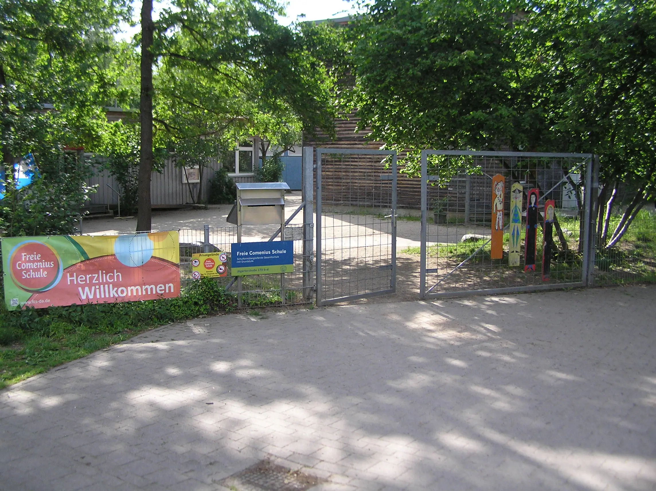 Ein Schuleingang mit einem Metalltor, bunten Schildern und einem Banner mit der Aufschrift „Herzlich Willkommen“. Hinter dem Tor sind an einem sonnigen Tag Bäume und ein Gebäude zu sehen.