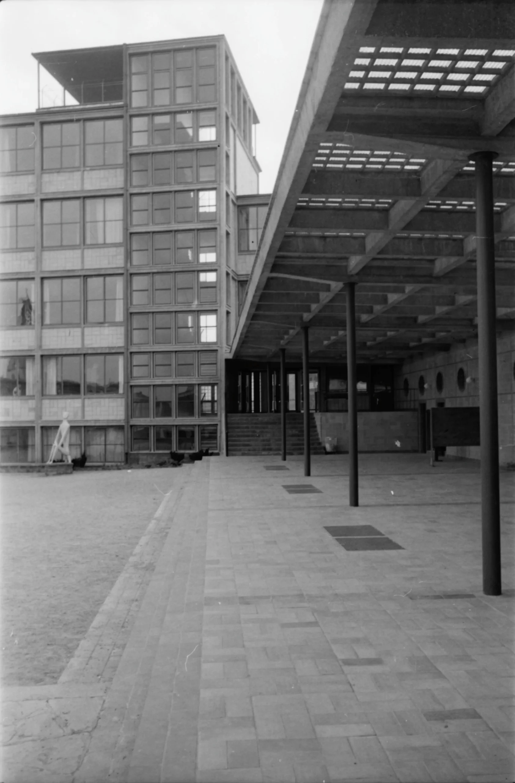 Schwarz-Weiß-Foto eines modernistischen Gebäudes mit Glasfenstern und einem von Säulen getragenen überdachten Gang. Der Bereich ist offen, gepflastert und größtenteils leer.