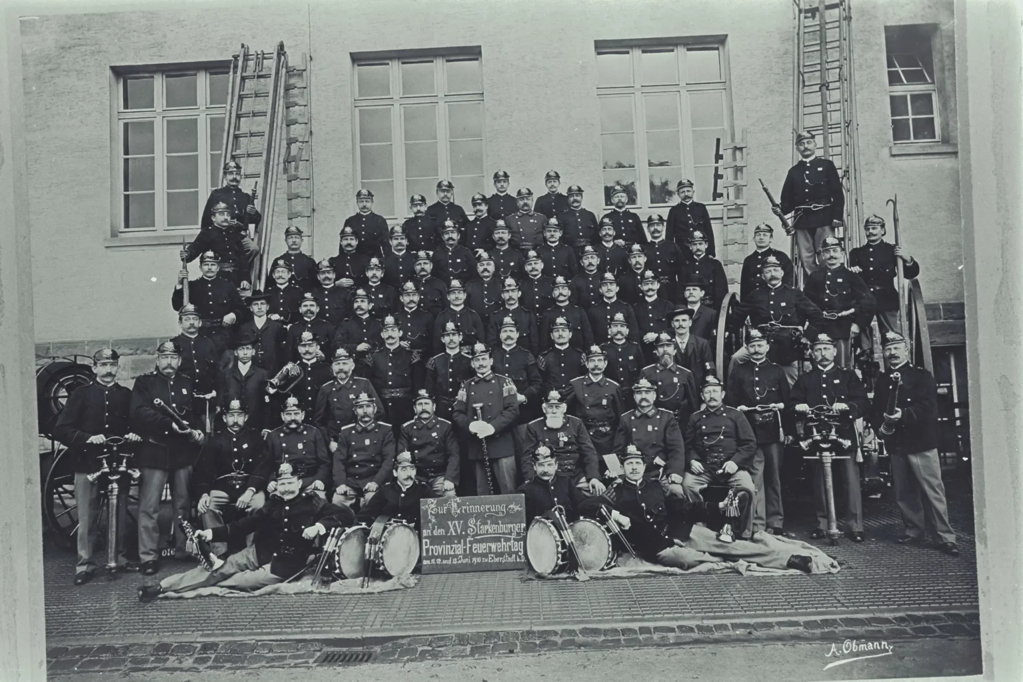 Ein Schwarz-Weiß-Foto, auf dem eine große Gruppe uniformierter Männer auf einer Treppe und vor einem Gebäude posiert; hinter ihnen stehen Leitern, davor liegen Musikinstrumente wie Trommeln und Hörner.