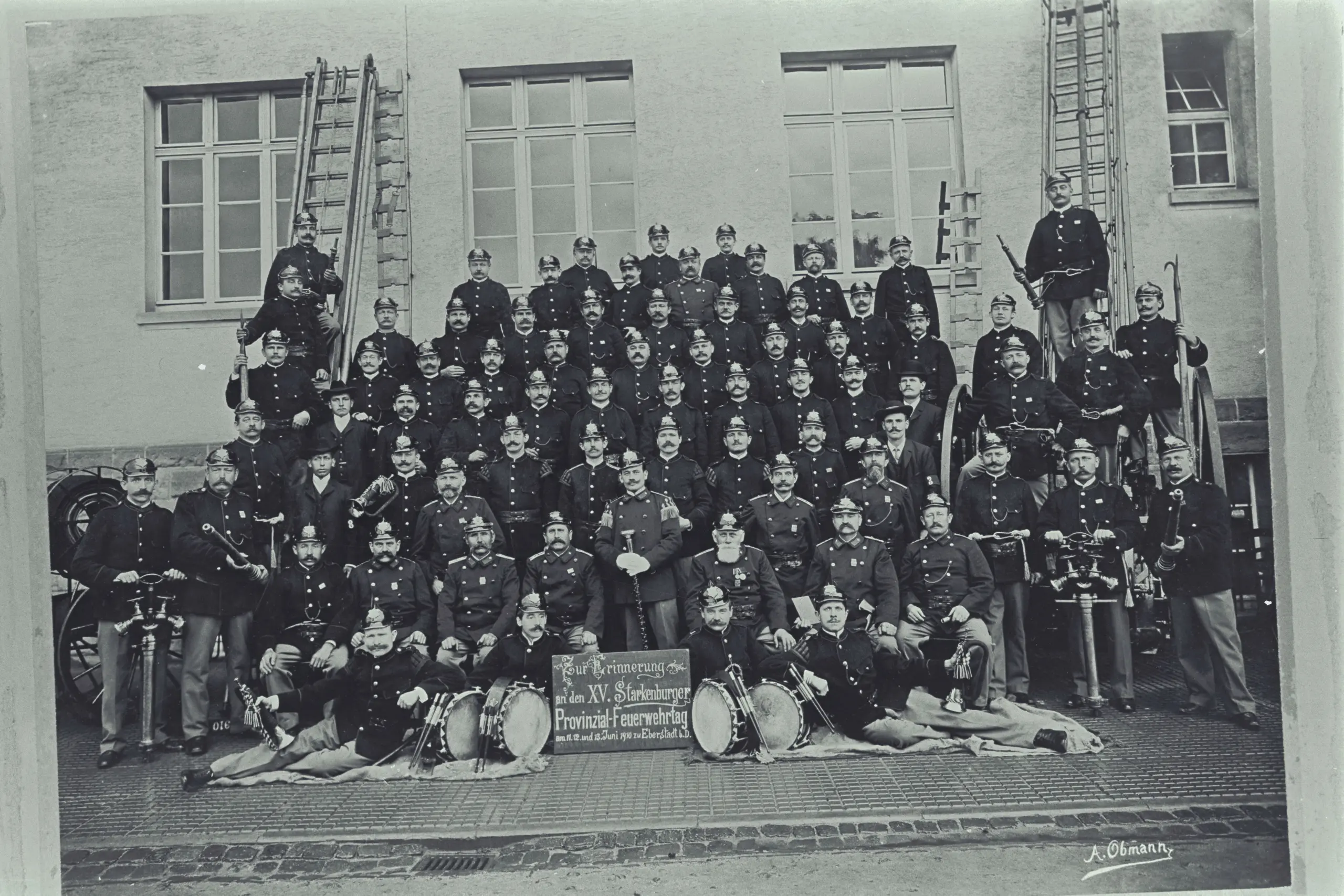 Ein Schwarz-Weiß-Foto, auf dem eine große Gruppe uniformierter Männer auf einer Treppe und vor einem Gebäude posiert; hinter ihnen stehen Leitern, davor liegen Musikinstrumente wie Trommeln und Hörner.