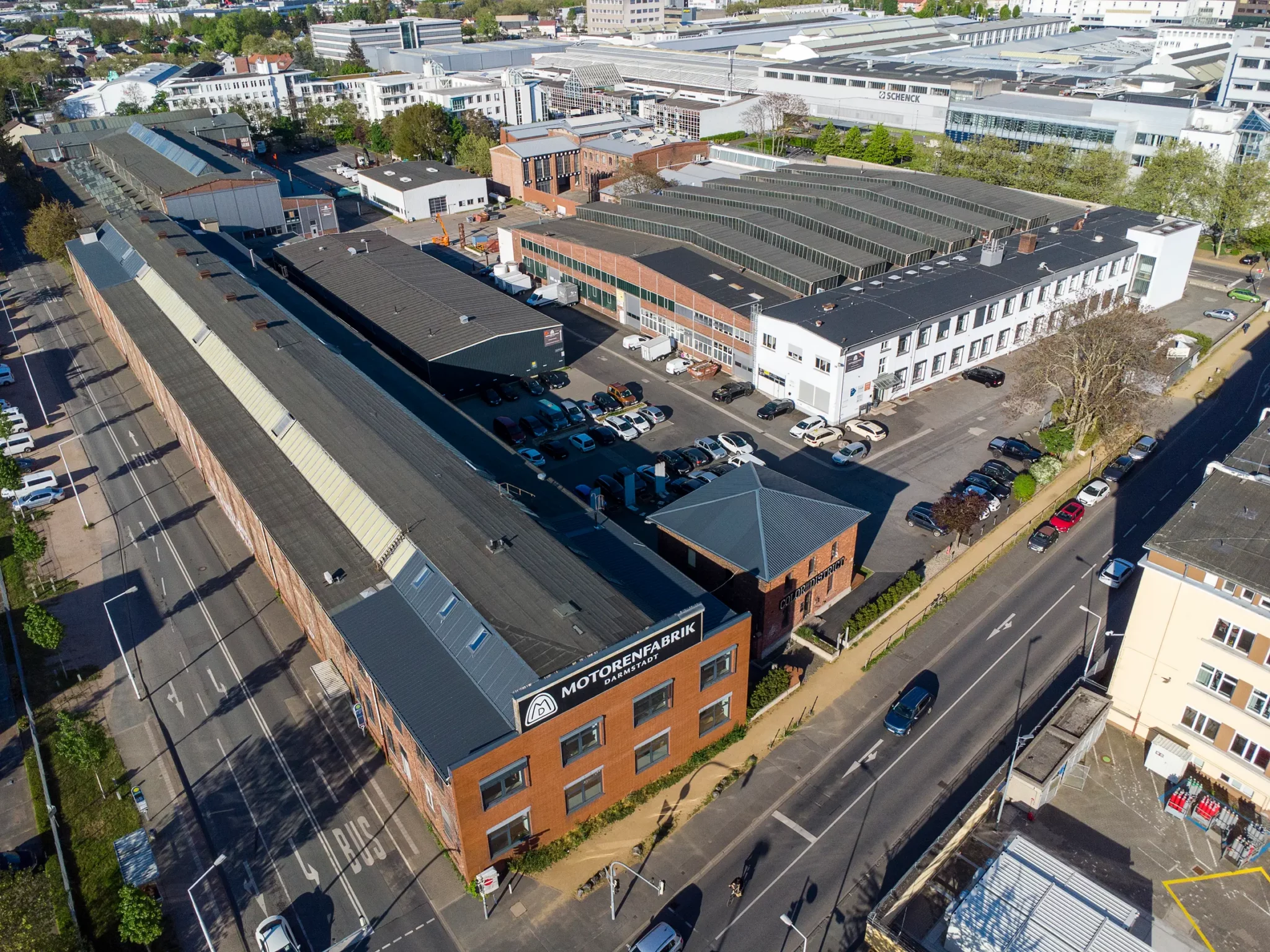 Luftaufnahme eines großen Industriekomplexes mit mehreren Gebäuden, Parkplätzen und angrenzenden Straßen. Am Hauptgebäude ist ein Schild mit der Aufschrift „Motopreneurpark“ zu sehen. Rund um die Gebäude stehen Autos, und die Straßen sind von Bäumen gesäumt.