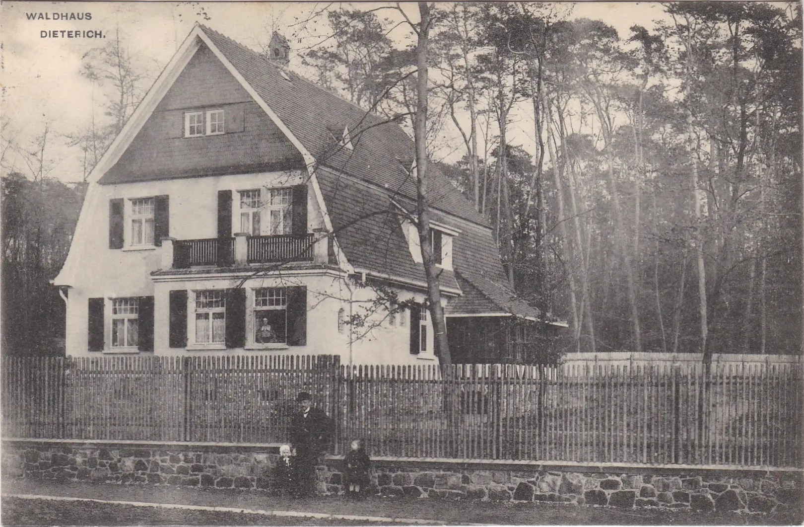 Schwarz-Weiß-Foto eines zweistöckigen Hauses mit steilem Dach, umgeben von einem Holzzaun und hohen Bäumen. Ein Mann und zwei Kinder sitzen auf dem Steinzaun davor. In der Ecke steht „Waldhaus Dieterich“.