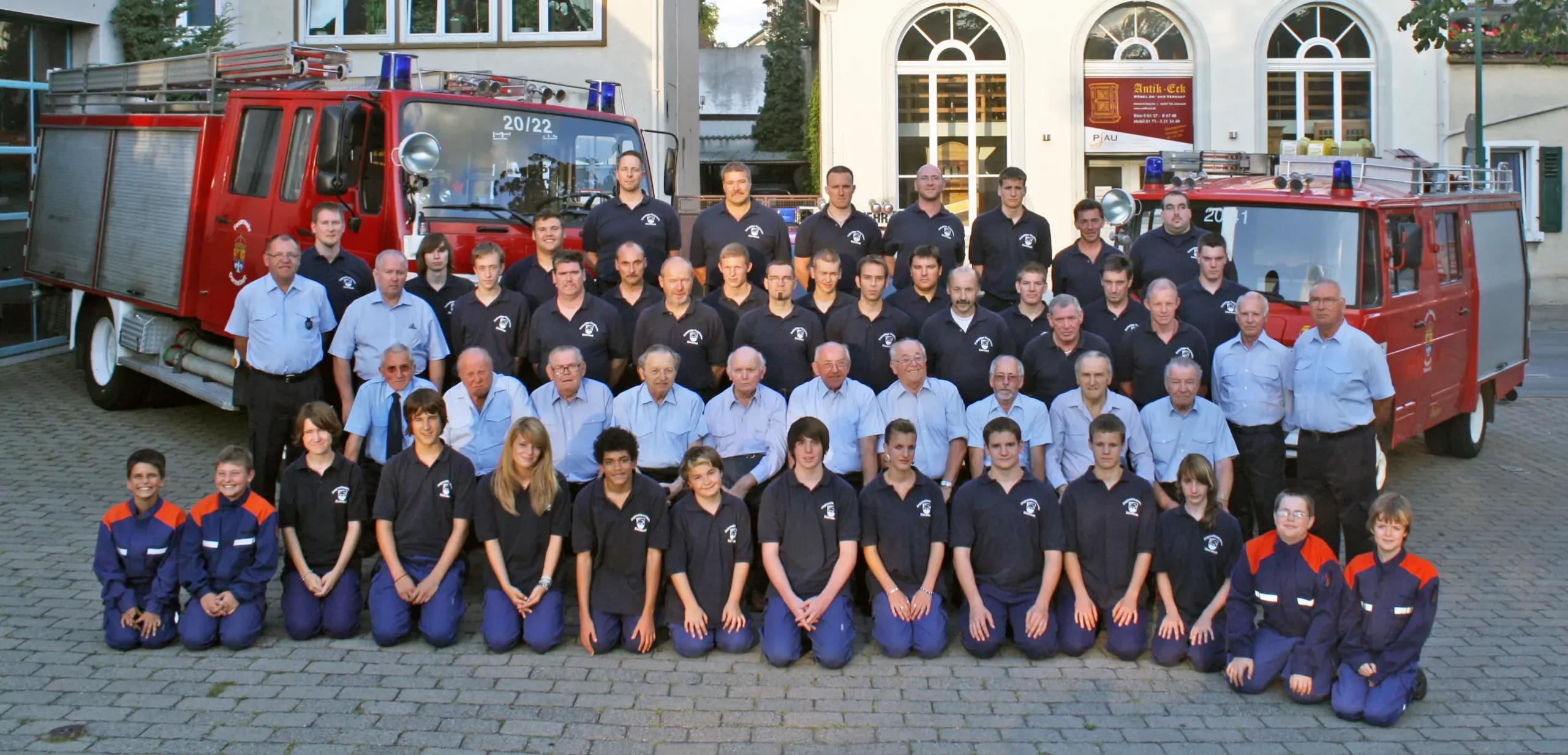 Eine große Gruppe von Feuerwehrleuten und Jugendkadetten posiert vor einer Feuerwache in Uniform für ein Gruppenfoto vor zwei roten Feuerwehrautos.