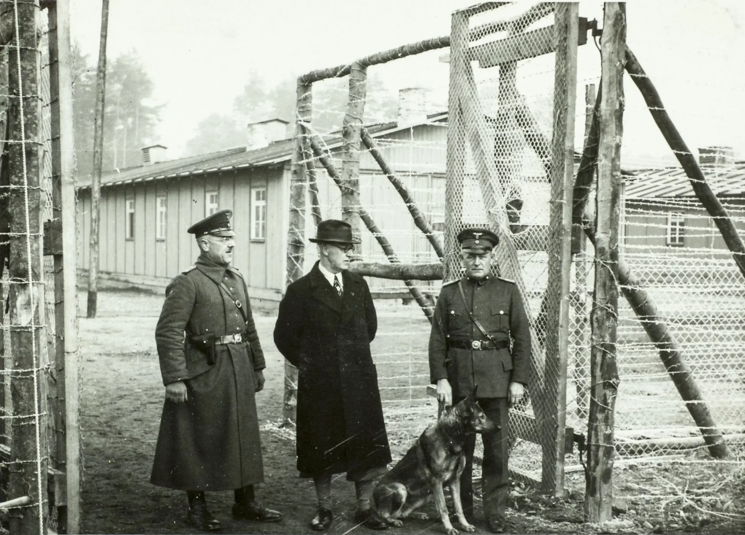 Drei Männer, zwei in Militäruniformen und einer in einem dunklen Mantel und mit Hut, stehen neben einem Stacheldrahtzaun und einem Tor. Zu ihren Füßen sitzt ein Deutscher Schäferhund. Im Hintergrund sind Holzgebäude und Bäume zu sehen.