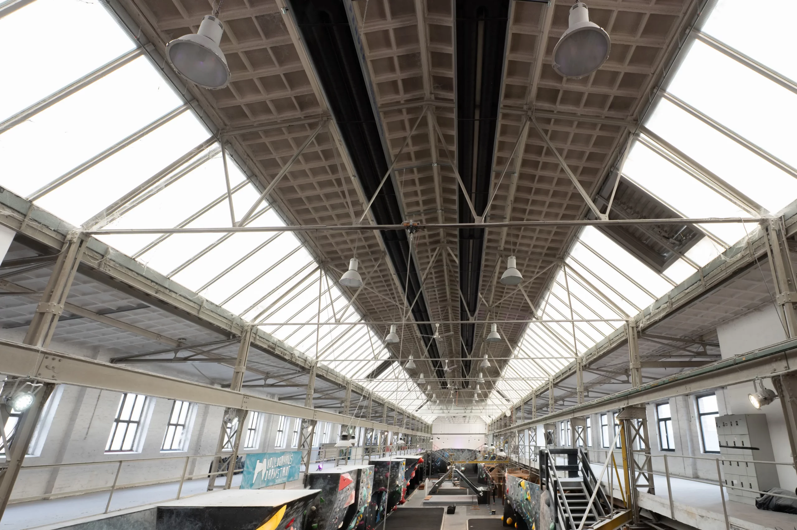 Innenansicht eines großen Industriegebäudes mit hoher, spitz zulaufender Decke, Oberlichtern, Leuchtstoffröhren, weißen Wänden und mehreren bunten Kletterwänden sowie Treppen, die im unteren Bereich zu sehen sind.