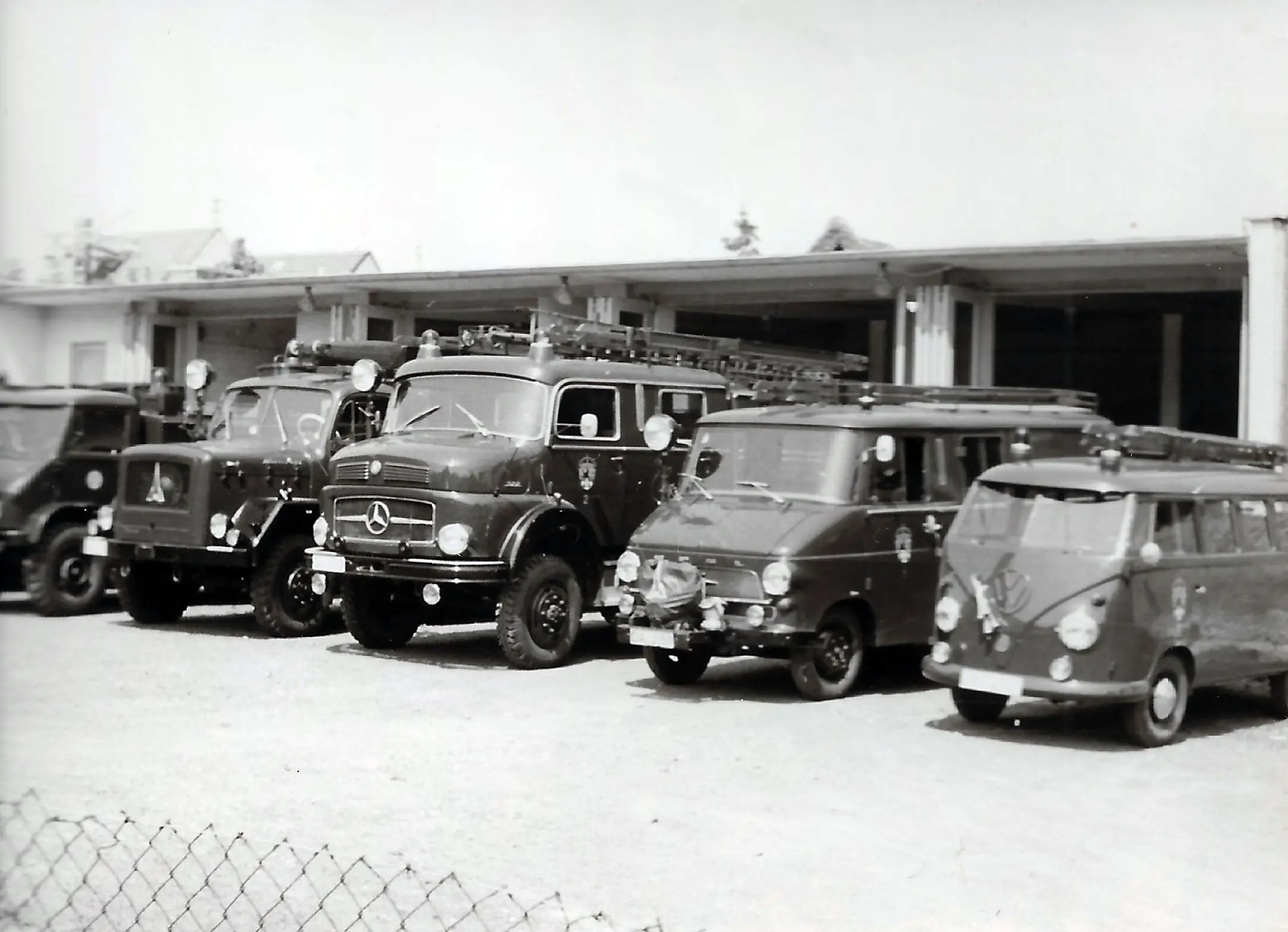 Vor einer offenen Garage stehen vier historische Feuerwehrfahrzeuge, darunter ein Mercedes-Benz und ein Volkswagen-Transporter. Das Bild ist in Schwarz-Weiß gehalten, und auf den Dächern der Fahrzeuge ist Feuerwehrausrüstung zu sehen.