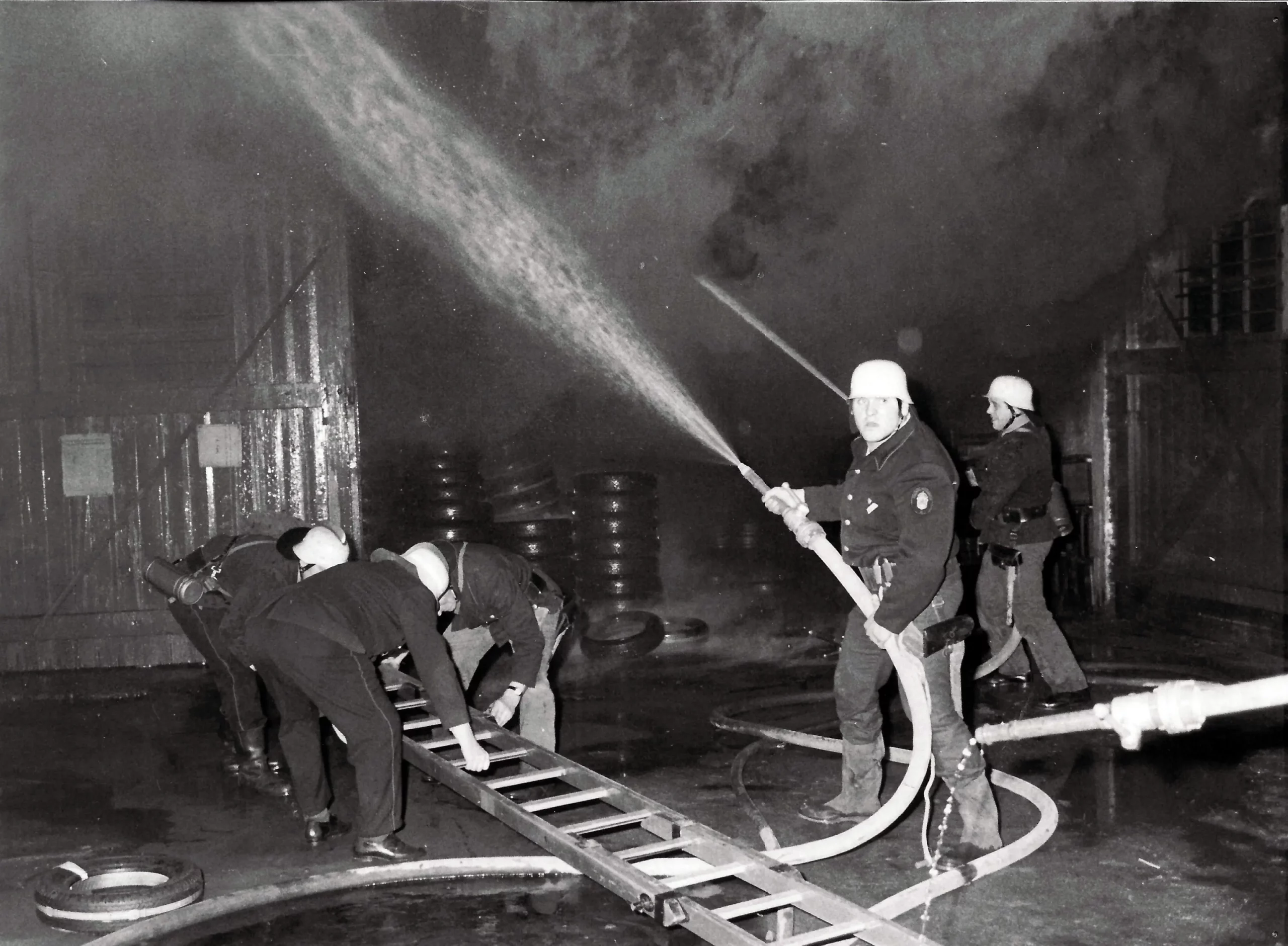 Feuerwehrleute mit Helmen spritzen Wasser und bedienen einen Schlauch, während andere eine Leiter auf einem nassen, rauchverhangenen Untergrund im Inneren eines Gebäudes aufstellen – vermutlich während eines Brandes.