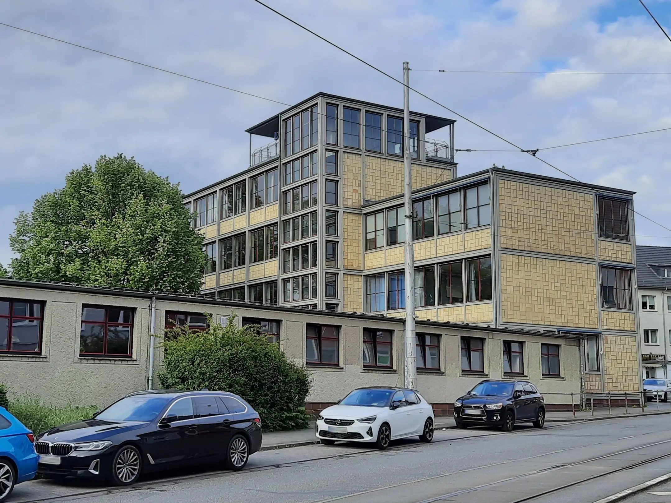 Ein modernes, mehrstöckiges Bürogebäude mit großen Fenstern und einer beigen Fassade, umgeben von geparkten Autos und Grünflächen unter einem teilweise bewölkten Himmel.