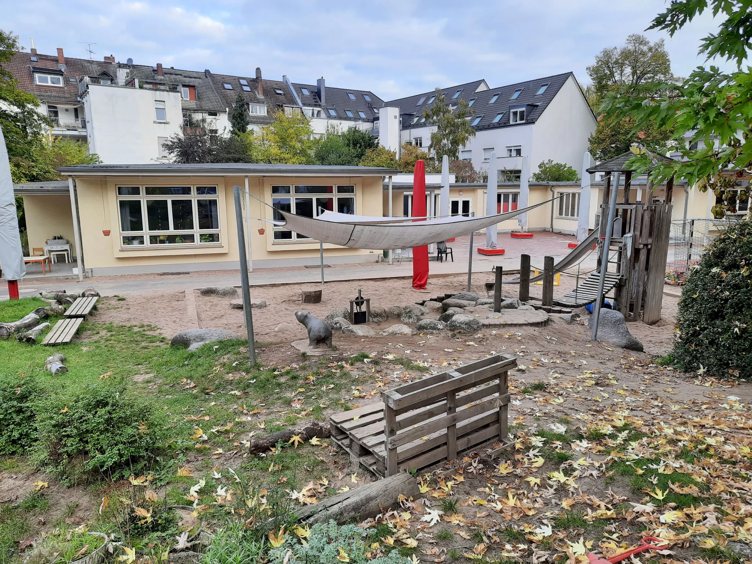 Ein Spielplatz mit Sand, Klettergerüsten aus Holz, einem Sonnendach und Bänken. Der Bereich ist von Rasen und Laub umgeben. Im Hintergrund sind gelbe Gebäude und Wohnhäuser zu sehen.