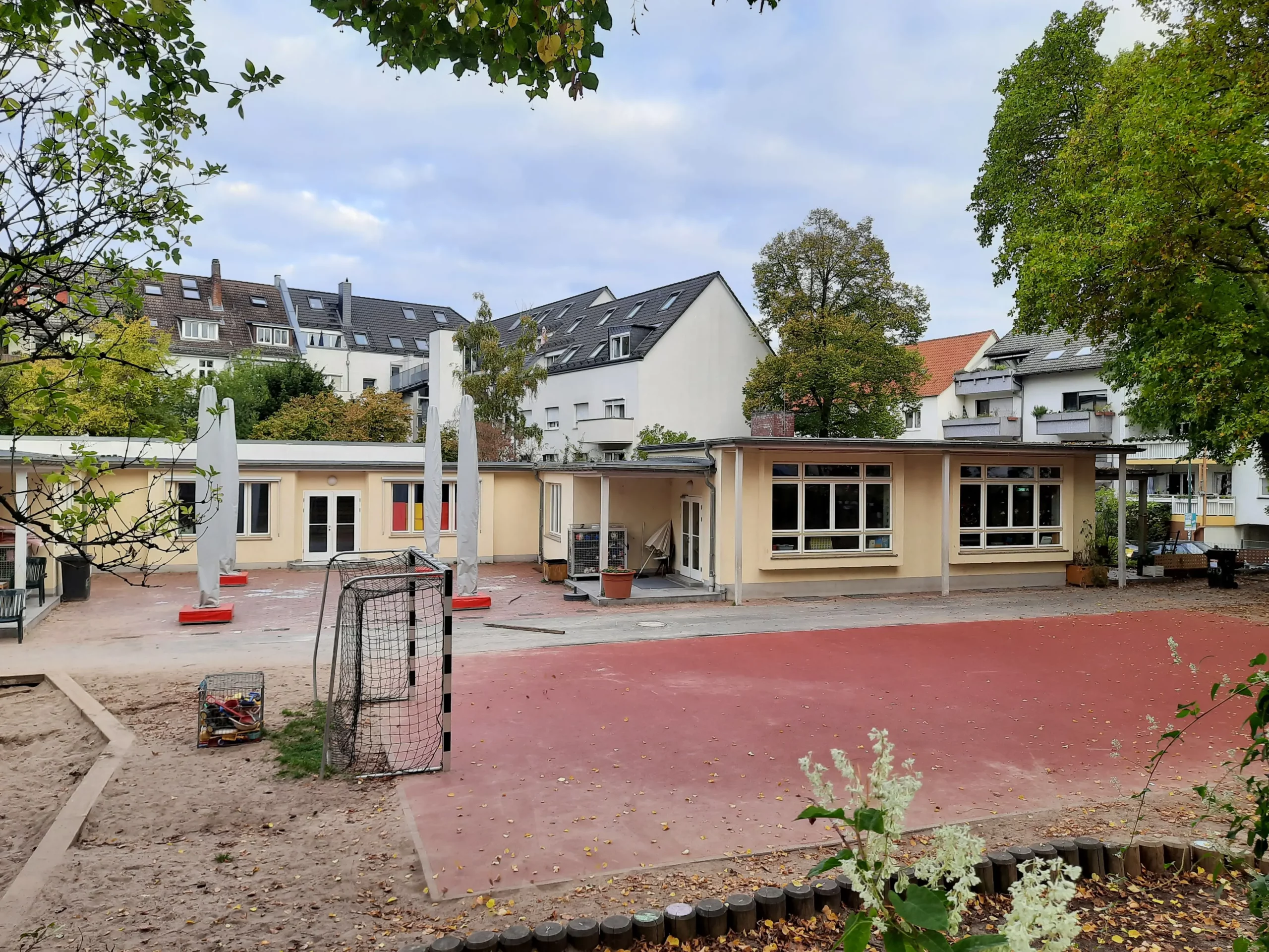 Ein Schulhof mit einem roten Sportplatz, einem kleinen Fußballtor, einer Sandfläche und einem niedrigen gelben Schulgebäude. Im Hintergrund sind Bäume und Wohnhäuser vor einem teilweise bewölkten Himmel zu sehen.