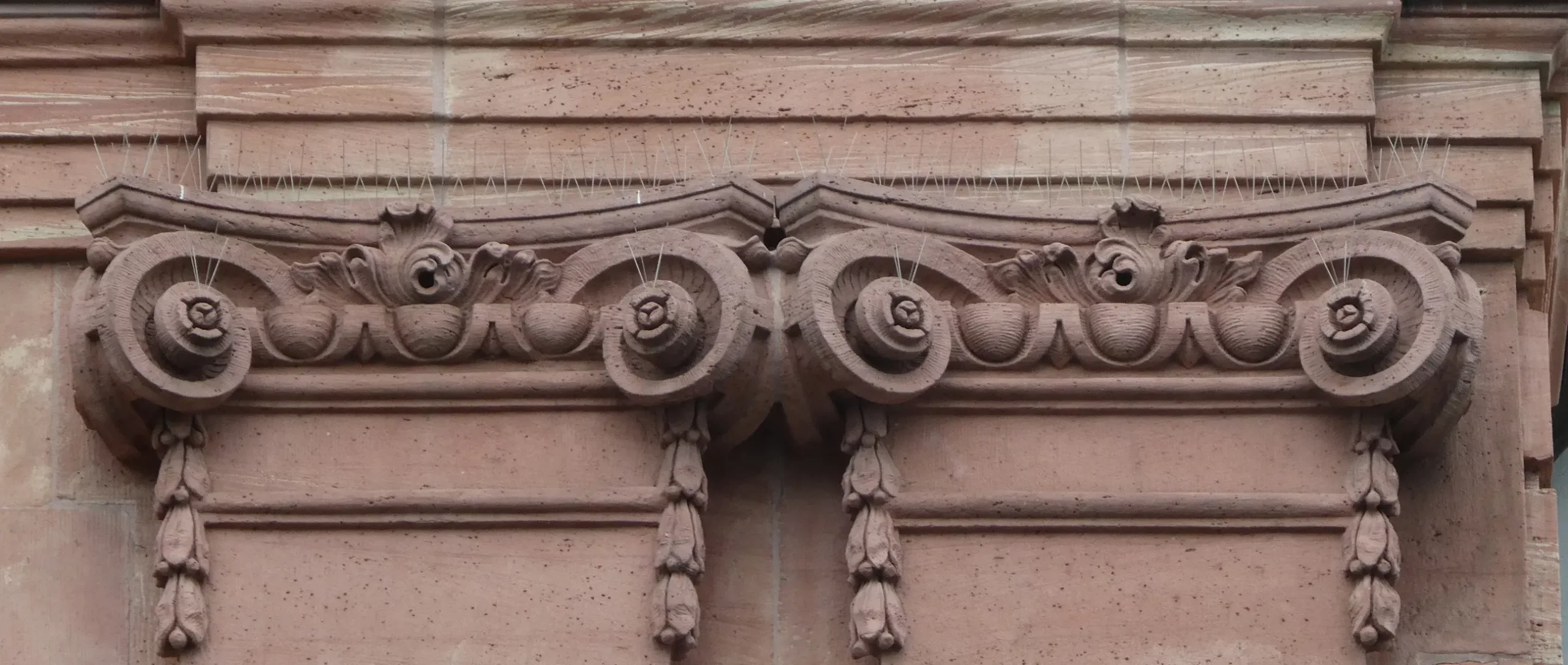 Aufwendig gearbeitetes architektonisches Steinrelief mit symmetrischen Blumen- und Spiralmotiven; die Oberfläche weist Verwitterungsspuren auf, und oben sind dünne Metallspitzen zu sehen, die Vögel abschrecken sollen.