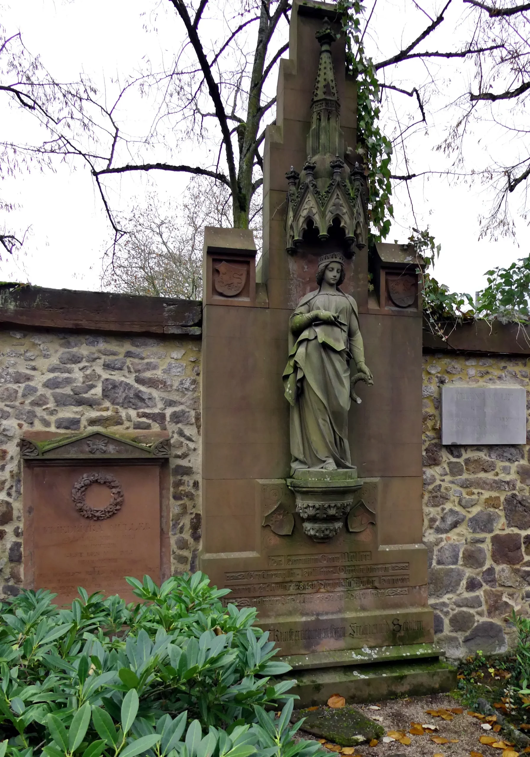 Ein Grabdenkmal aus Stein mit der Statue einer Frau in einem wallenden Kleid, das vor einer alten Steinmauer steht und von grünen Sträuchern und kahlen Ästen umgeben ist.