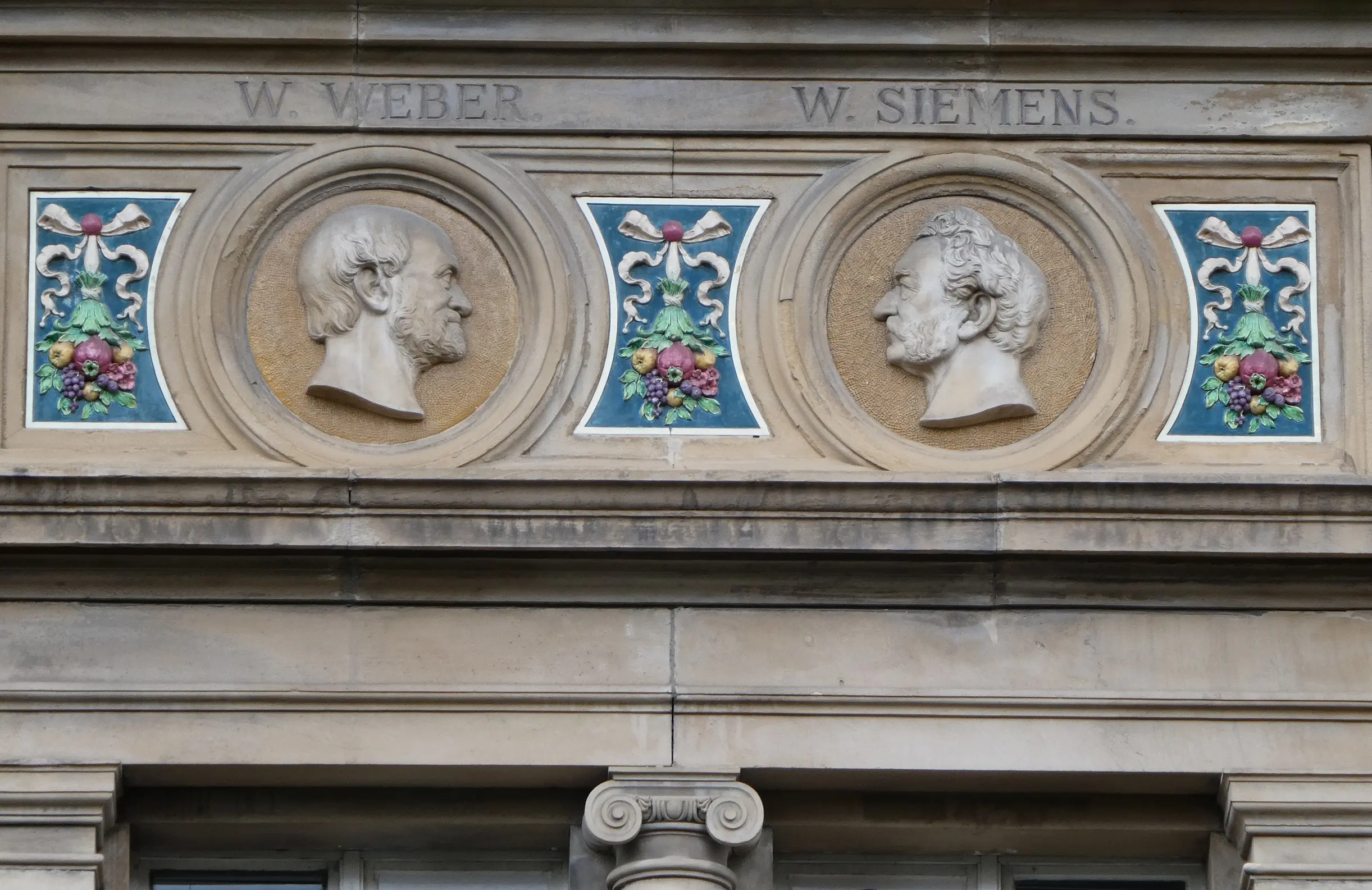 Fassade eines Steingebäudes mit zwei im Seitenprofil geformten Büsten mit den Aufschriften „W. Weber“ und „W. Siemens“, die durch farbenfrohe Blumenverzierungen über einem Fenster voneinander getrennt sind.