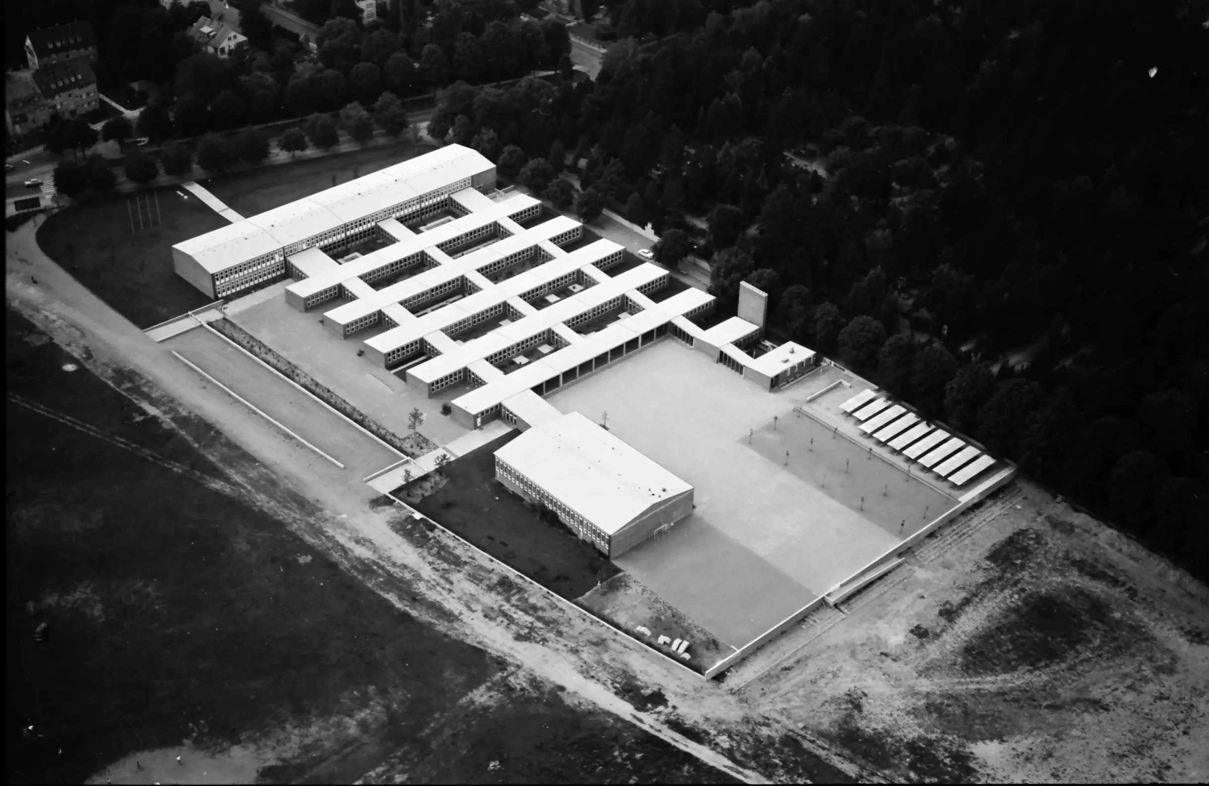 Luftaufnahme eines modernistischen Gebäudekomplexes mit rasterartigem Grundriss, umgeben von Bäumen und Freiflächen, mit rechteckigen weißen Gebäuden, offenen Innenhöfen und angrenzenden Sportplätzen.