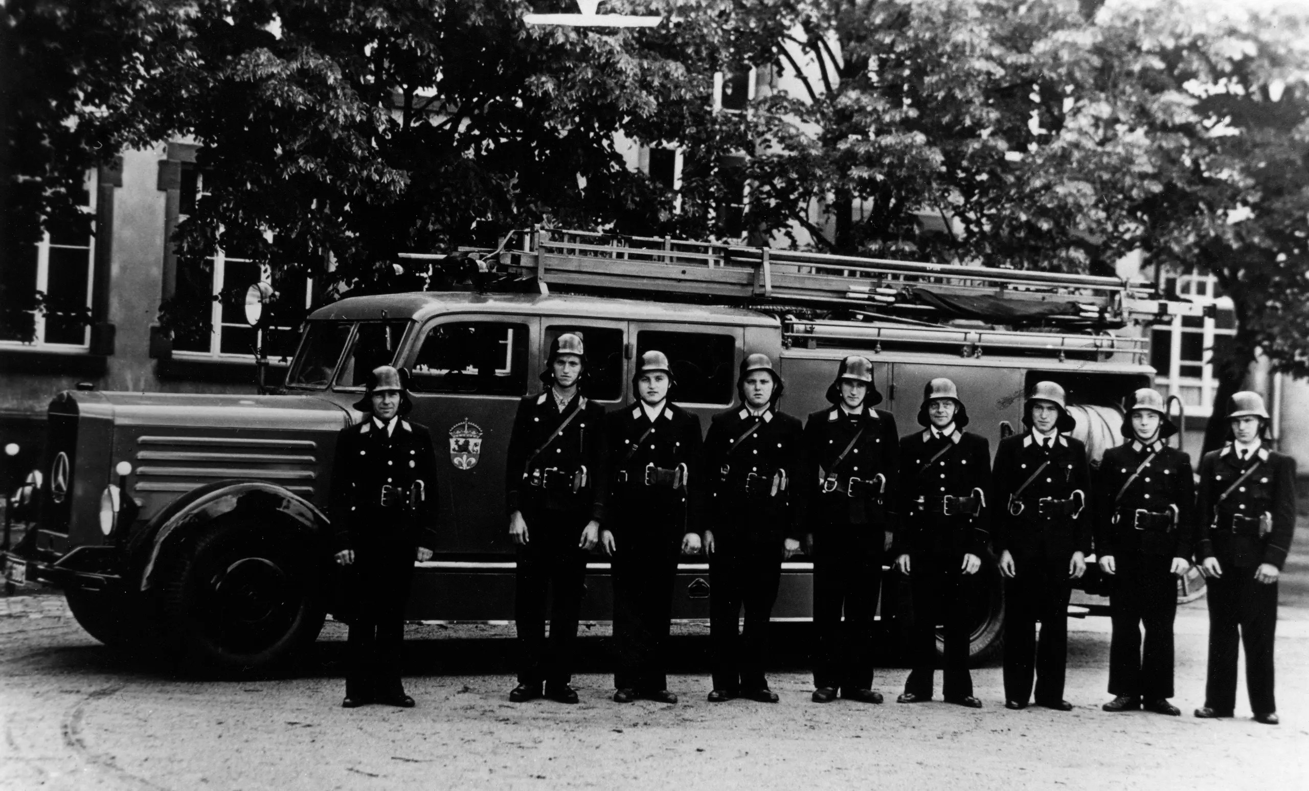 Neun Feuerwehrleute in Uniform stehen in einer Reihe vor einem alten Feuerwehrauto, im Hintergrund sind Bäume und ein Gebäude zu sehen. Das Bild scheint schwarz-weiß zu sein.