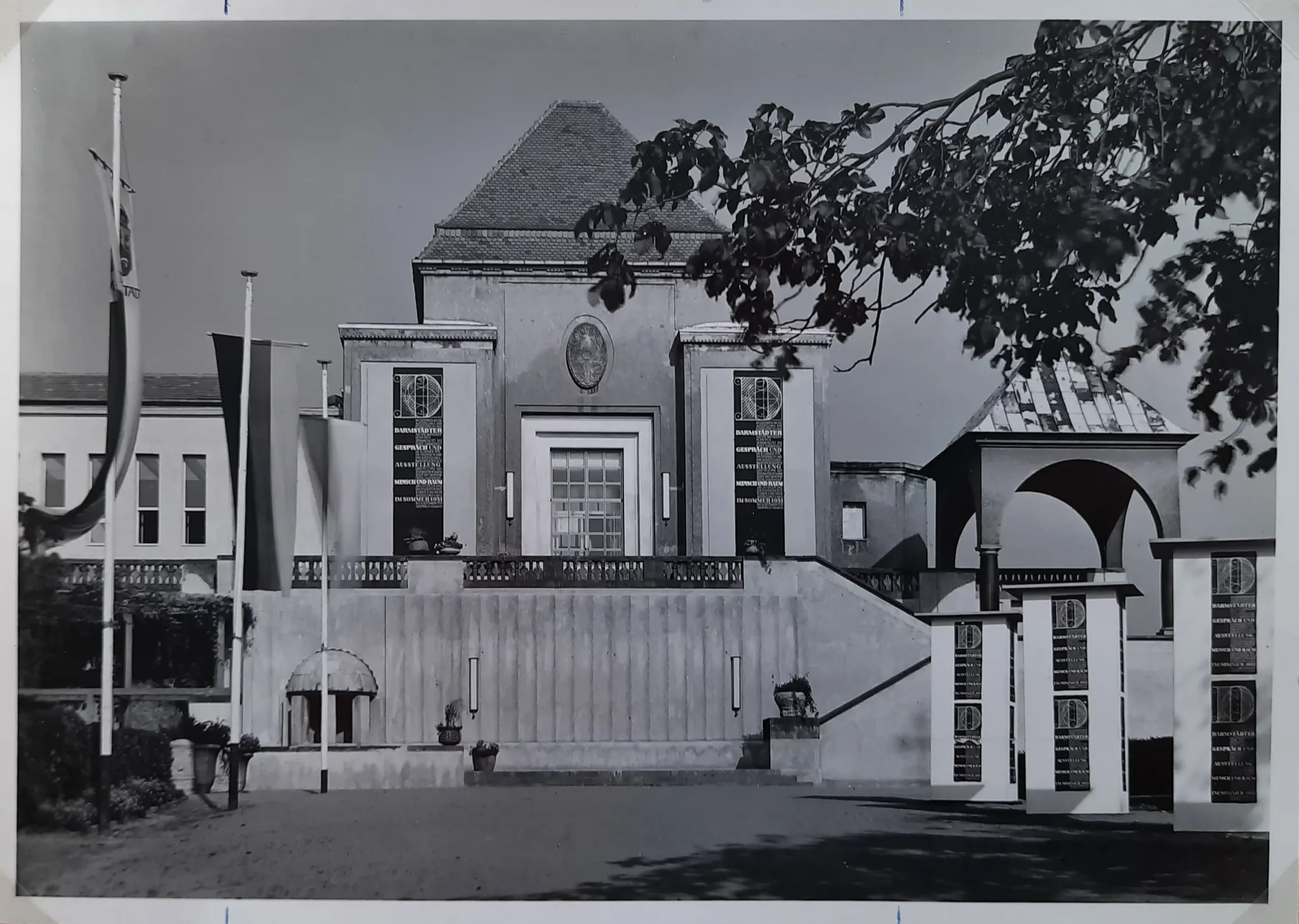 Schwarz-Weiß-Foto eines repräsentativen Gebäudes mit großen Fenstern, einem steilen Dach, Säulen und einer Treppe, die nach oben führt. Der Bereich oben rechts ist teilweise von Bäumen verdeckt. Auf der linken Seite sind Flaggen zu sehen.