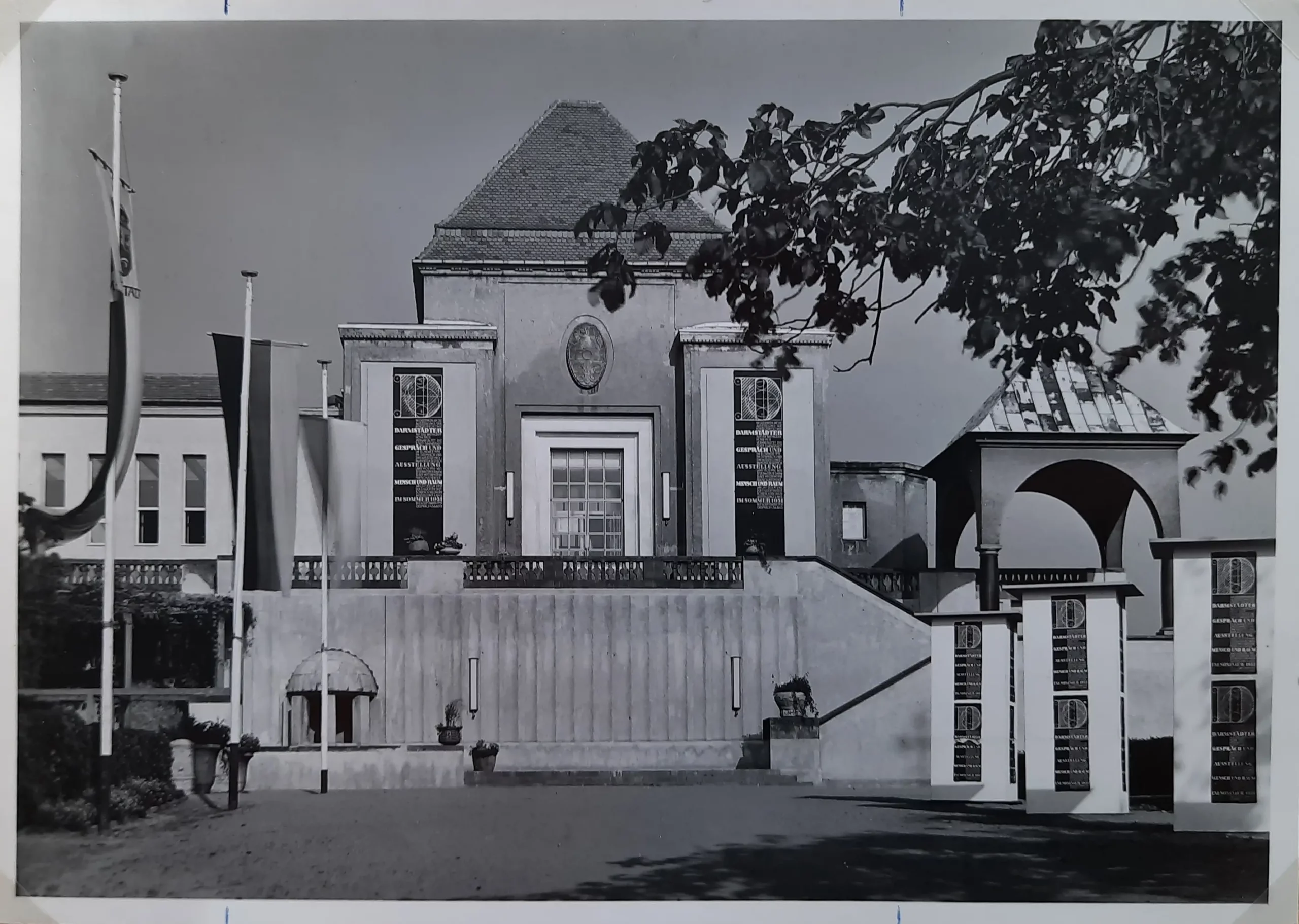 Schwarz-Weiß-Foto eines repräsentativen Gebäudes mit großen Fenstern, einem steilen Dach, Säulen und einer Treppe, die nach oben führt. Der Bereich oben rechts ist teilweise von Bäumen verdeckt. Auf der linken Seite sind Flaggen zu sehen.