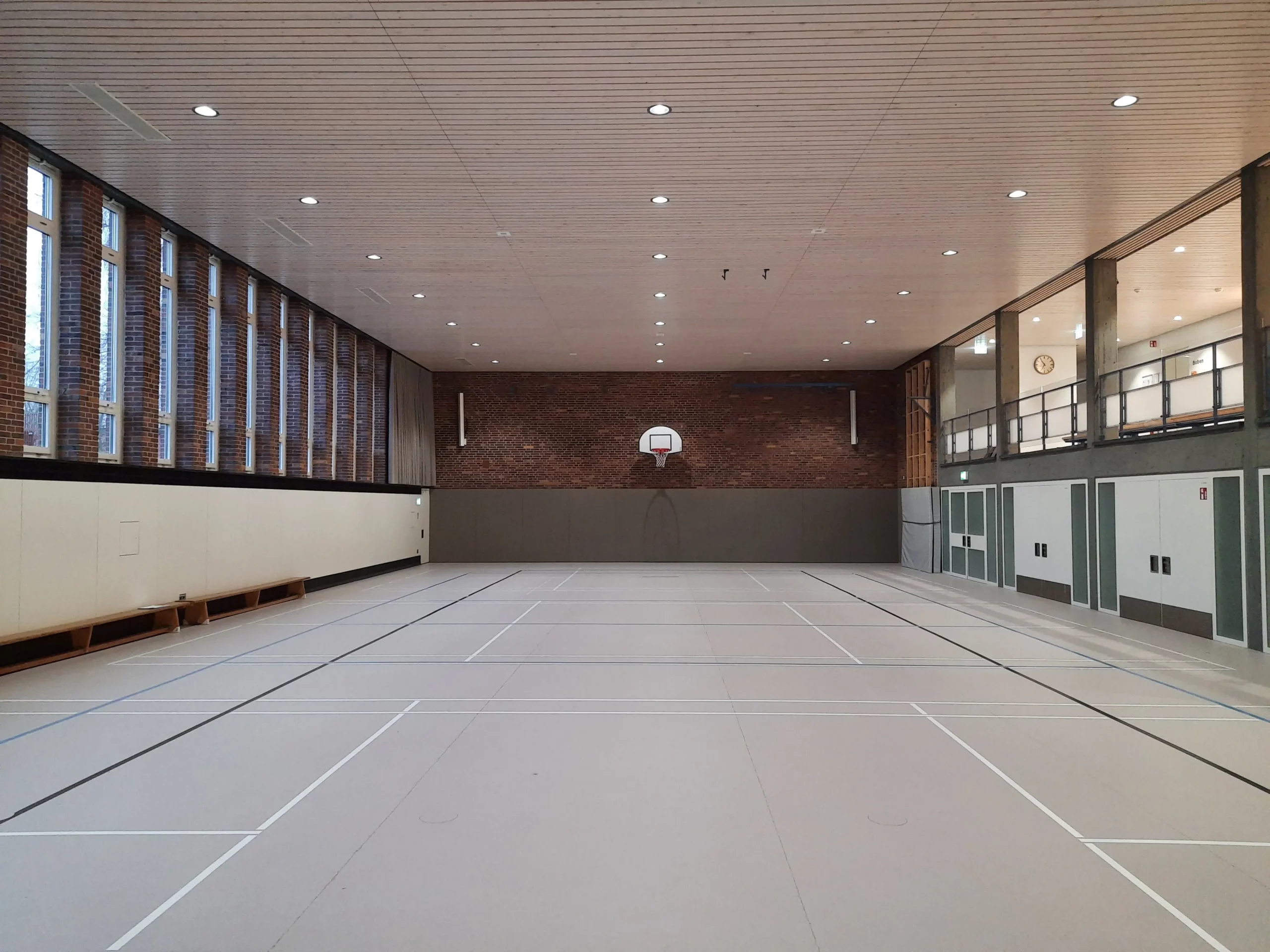 Eine leere Sporthalle mit einem Basketballkorb an der gegenüberliegenden Wand, einer Holzdecke, hohen Fenstern auf der linken Seite und einem Balkonbereich mit Geländer und Türen auf der rechten Seite. Weiße Begrenzungslinien markieren das helle Spielfeld.