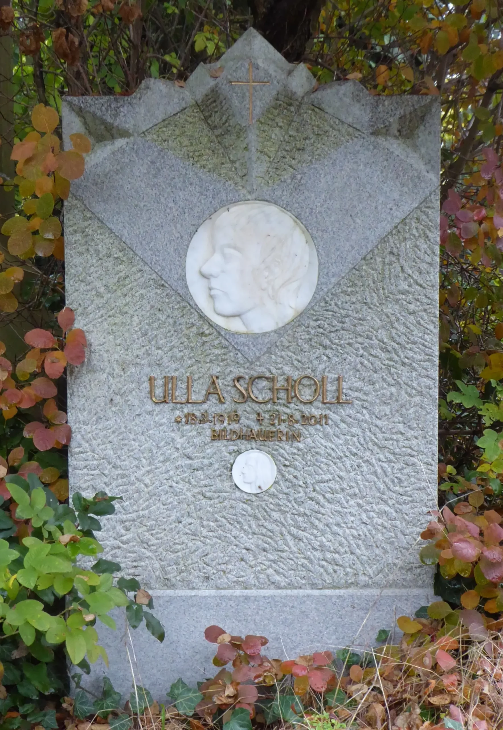 Ein grauer Grabstein mit einem eingravierten Profilporträt und der goldenen Inschrift „Ulla Scholl“, umgeben von grünem und rotem Laub.